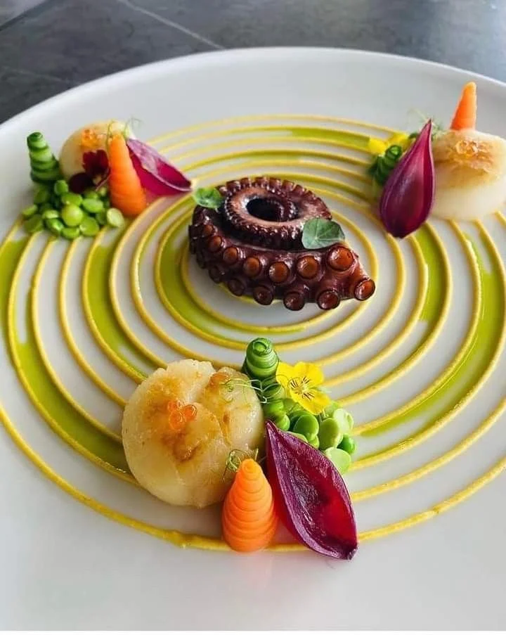 A gourmet dish featuring a grilled octopus tentacle at the center, surrounded by colorful vegetables and garnishes on a white plate with yellow sauce and concentric circles of green sauce.