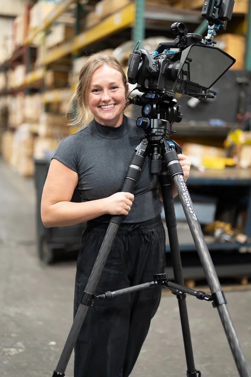 A woman with shoulder-length blonde hair smiling while holding a professional video camera on a tripod in a warehouse with shelves of boxes.