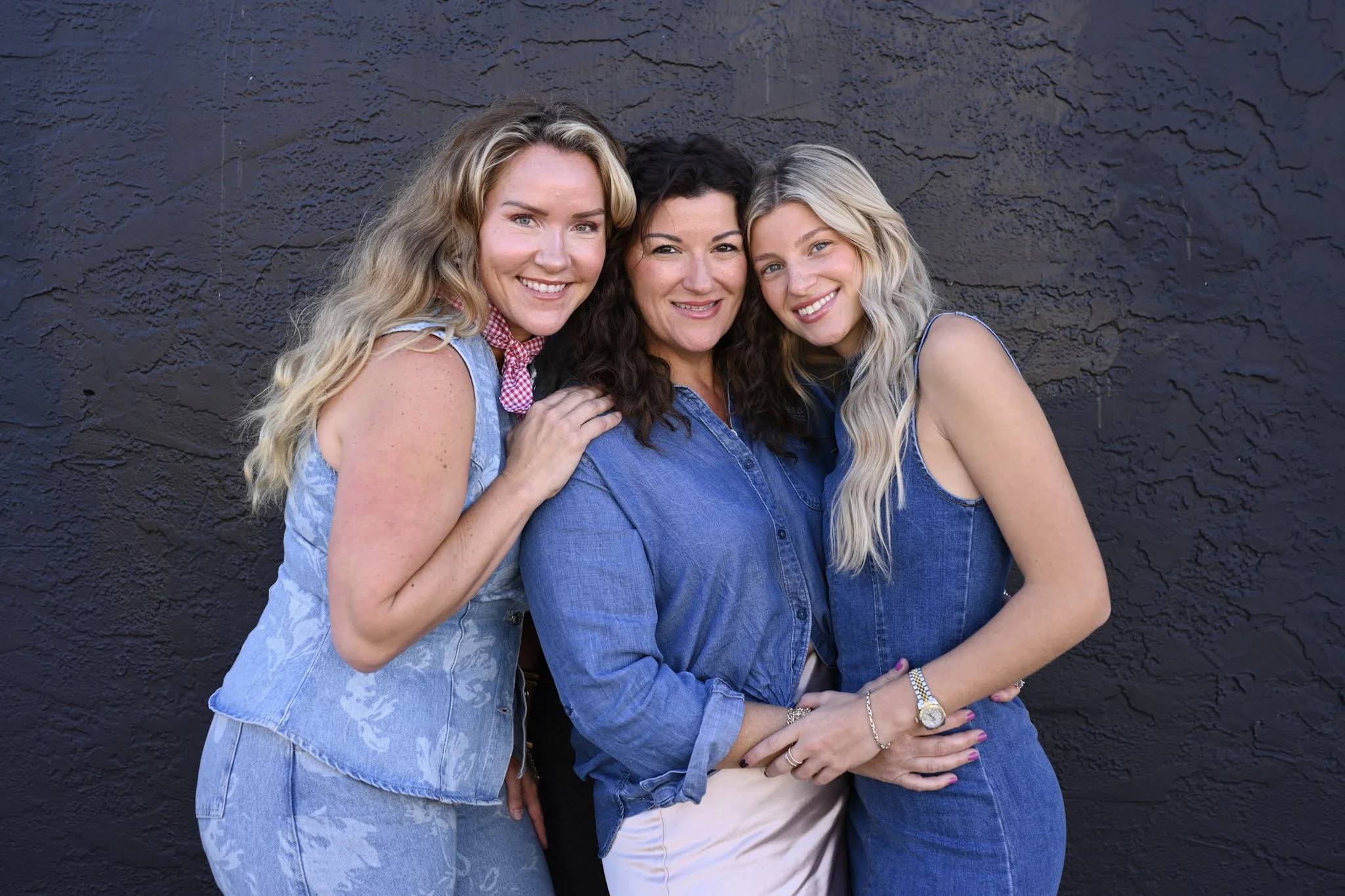 Three women standing close together against a black wall, smiling at the camera. All are wearing denim clothing.