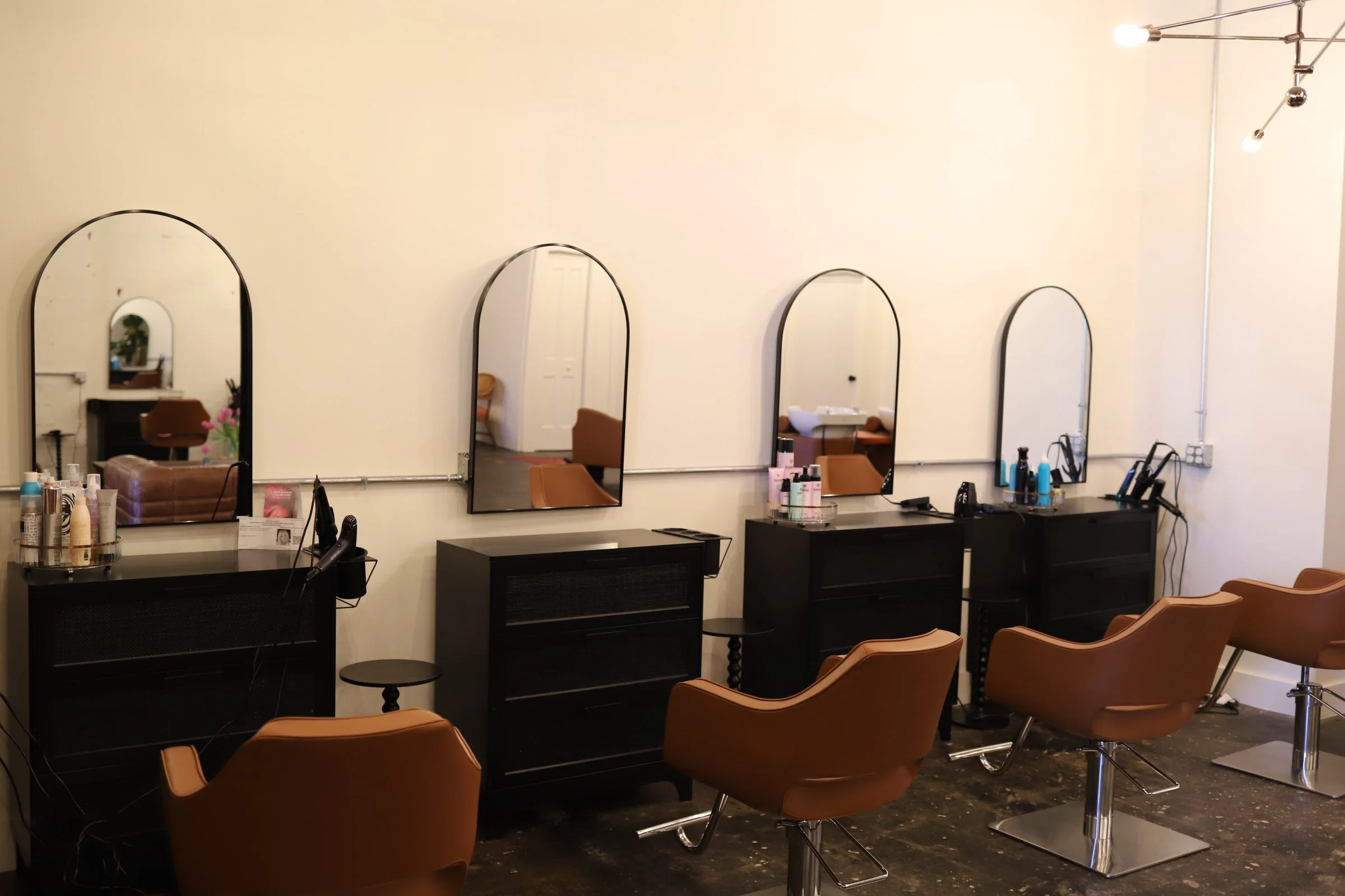 Hair salon with four hair styling stations, each with a mirror, hair products, and styling tools. Four brown salon chairs face the mirrors, and a modern light fixture hangs on the ceiling.