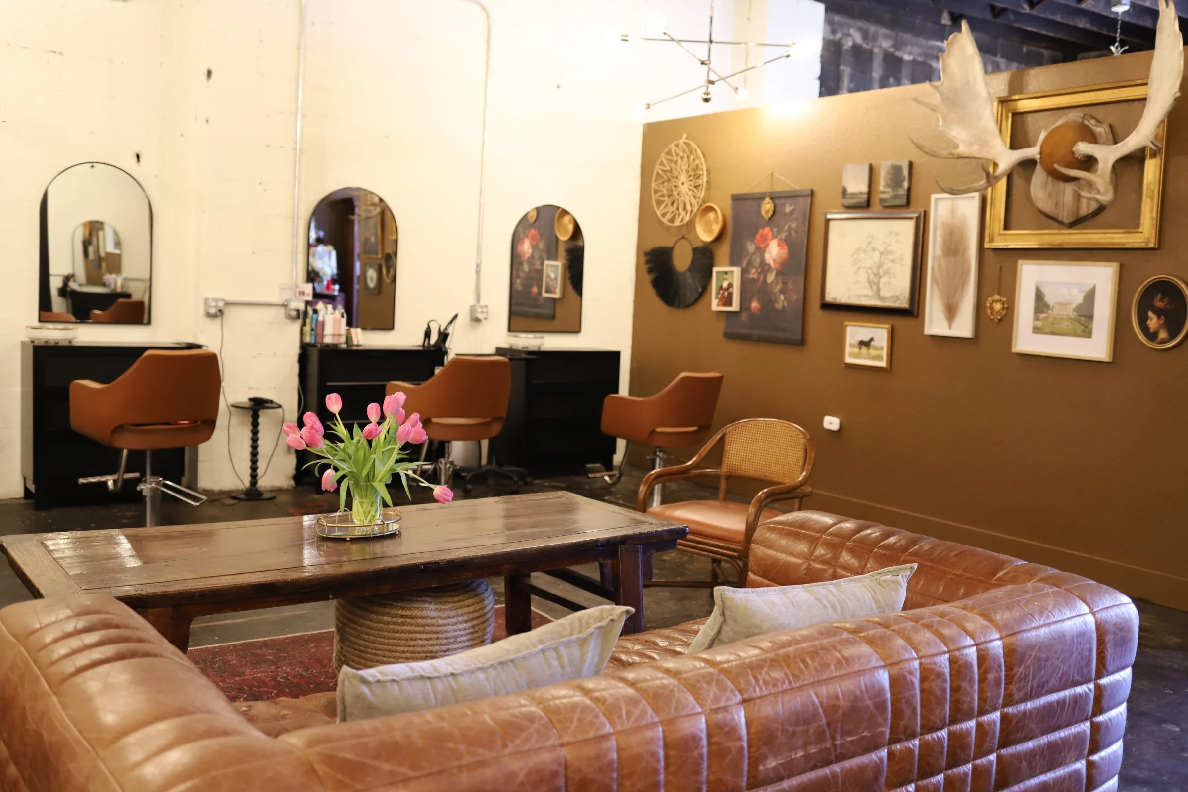 Interior of a stylish lounge or salon with a leather sofa, wooden coffee table, and decorated wall with framed artwork and a mounted moose head. There are two salon chairs and mirrors on the back wall.