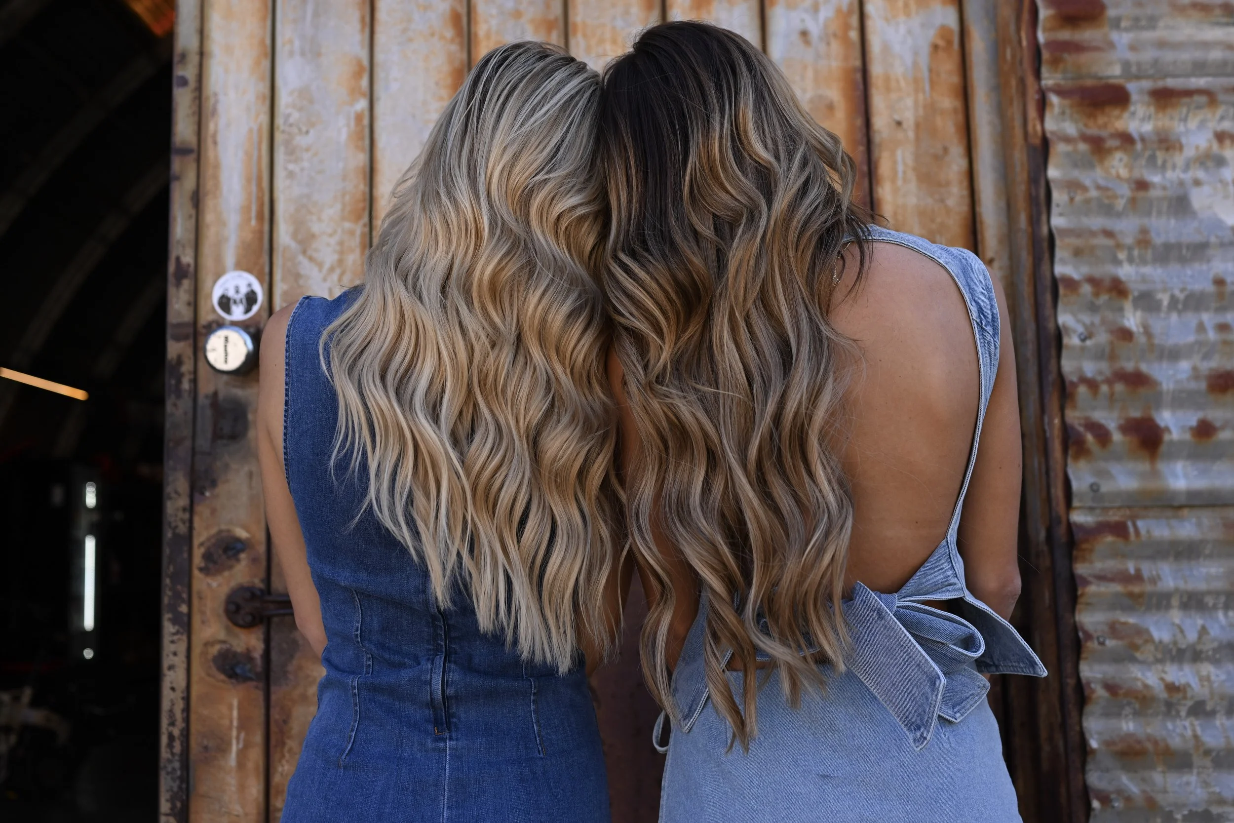 Two women with long, wavy blonde and brown hair, wearing denim dresses, leaning their foreheads together against a rustic wooden background.