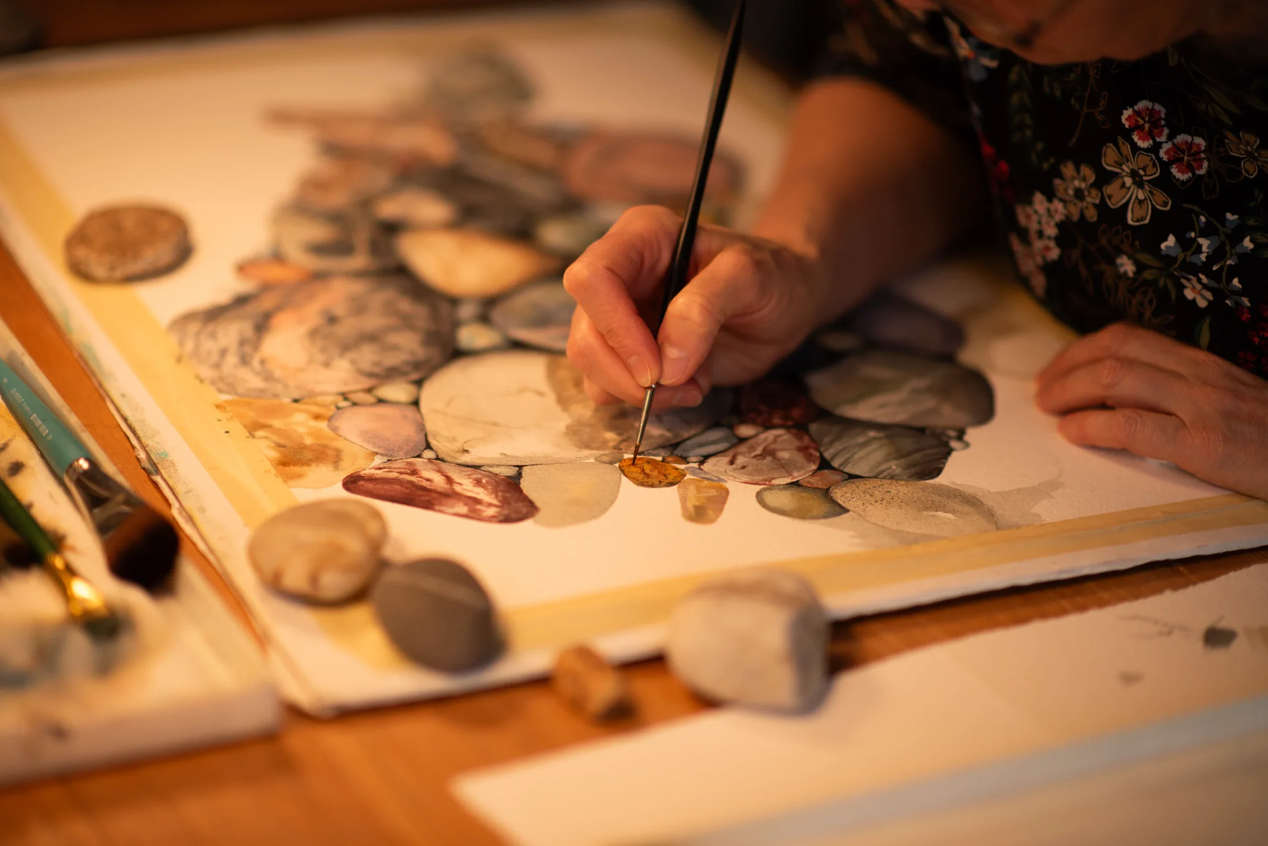 A person with painted fingernails using a fine brush to paint details on a watercolor painting of rocks and shells.