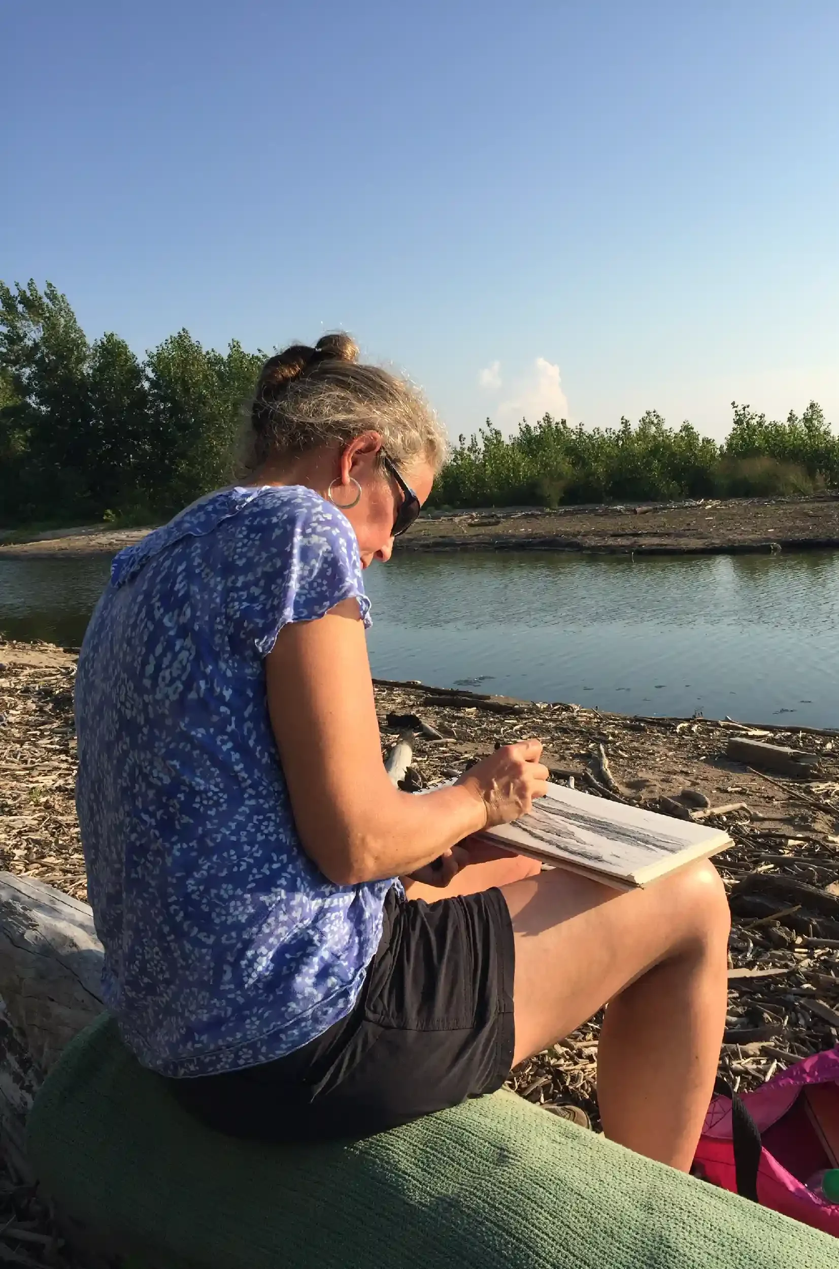 A woman sitting on a log by the water, reading a book, with green trees in the background and a clear blue sky.