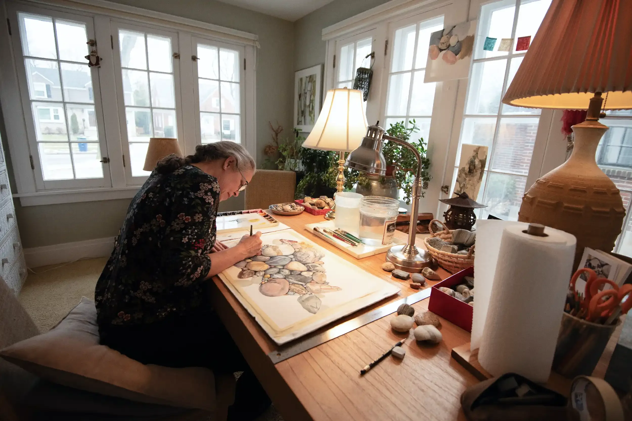 Wendy Caldwell Maloney hunched over a painting of stones. In the background are large windows. Her desk is covered with watercolor paints and accessories.