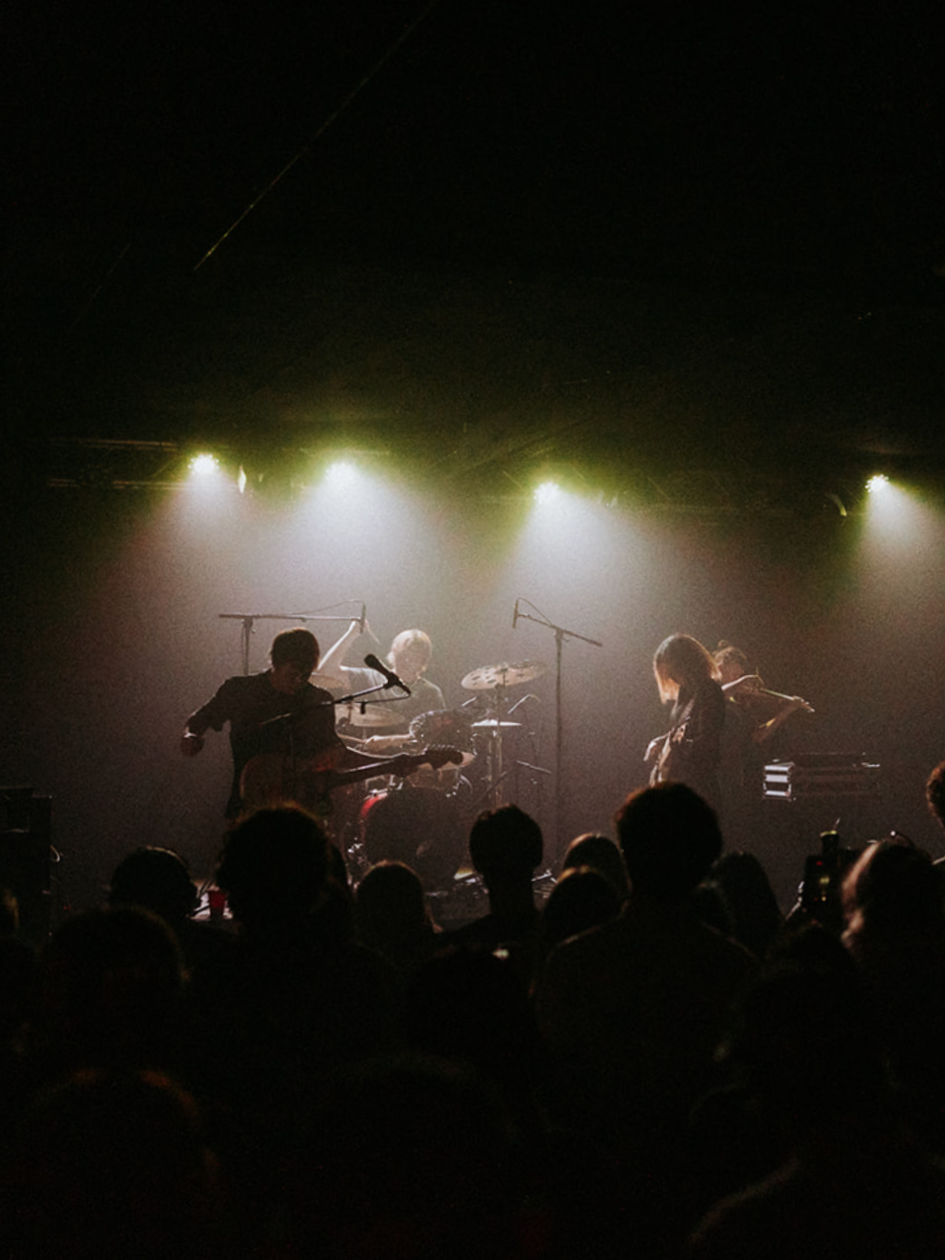 A band performing on stage with four members and audience silhouettes in the foreground, under bright stage lights.