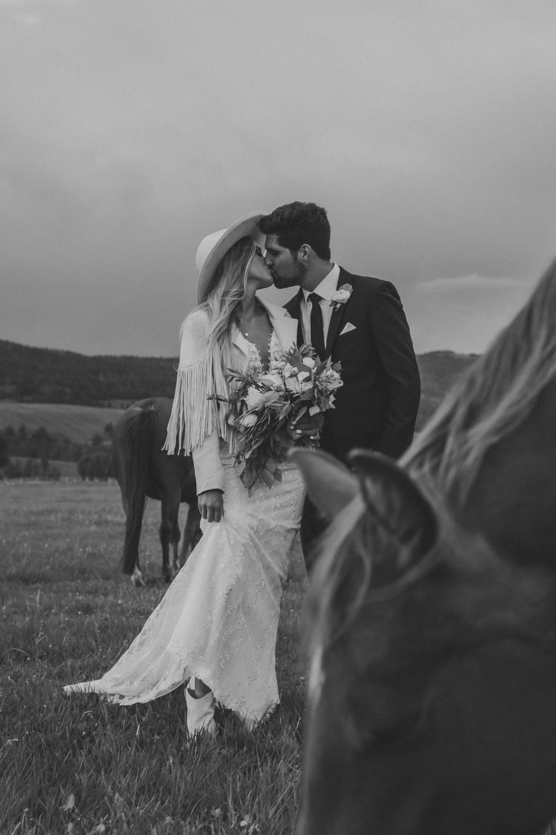 Black and white photo of a bride and groom kissing outdoors, with horses in the background.