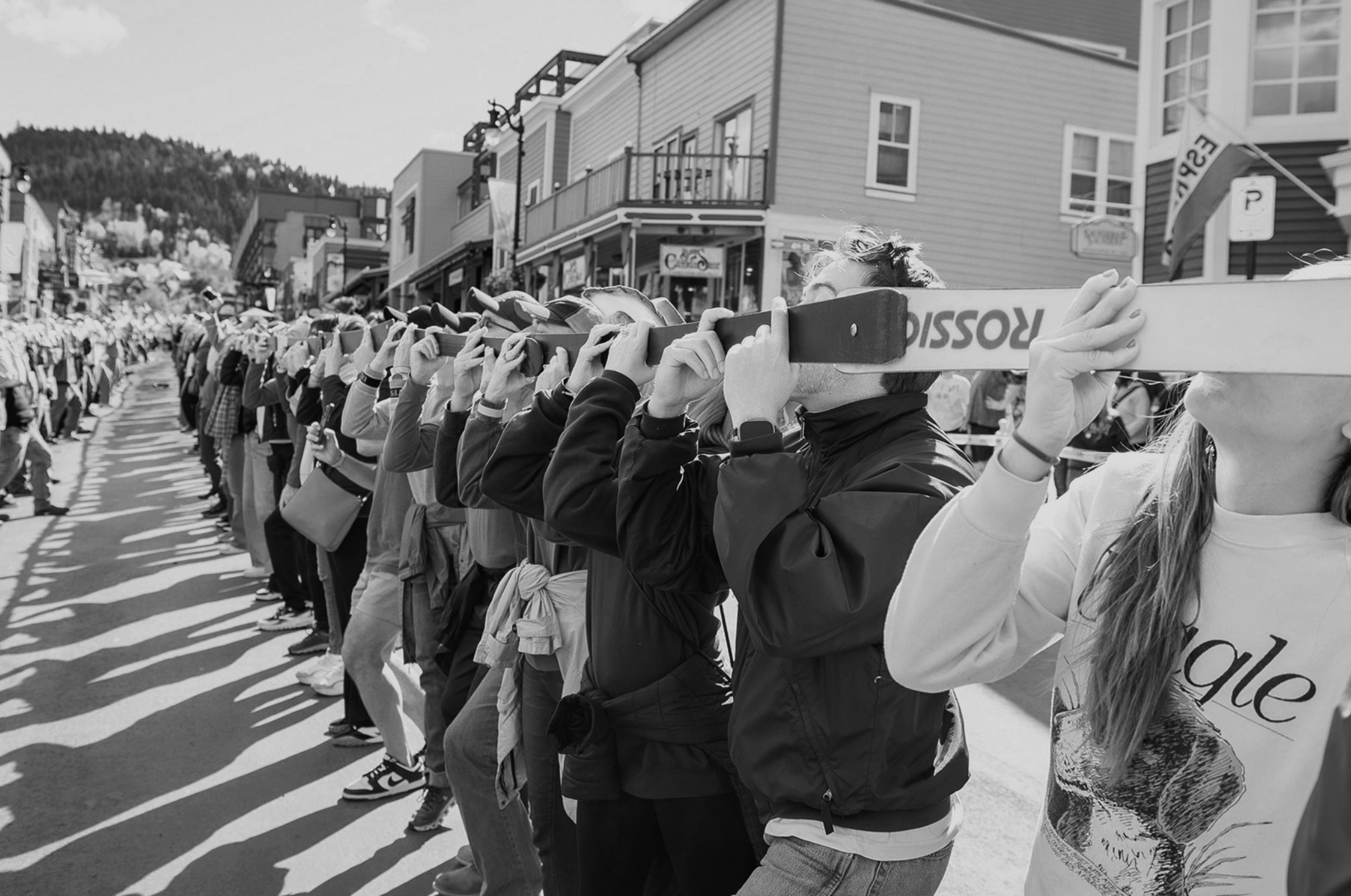 A line of people standing on a street, each person holding a shot-ski to their mouths, with a row of shops and buildings in the background. In Park City Utah
