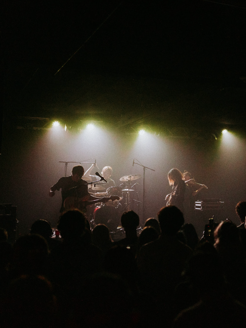 A live music concert with a band performing on stage under bright yellow and white spotlights, and an audience watching.