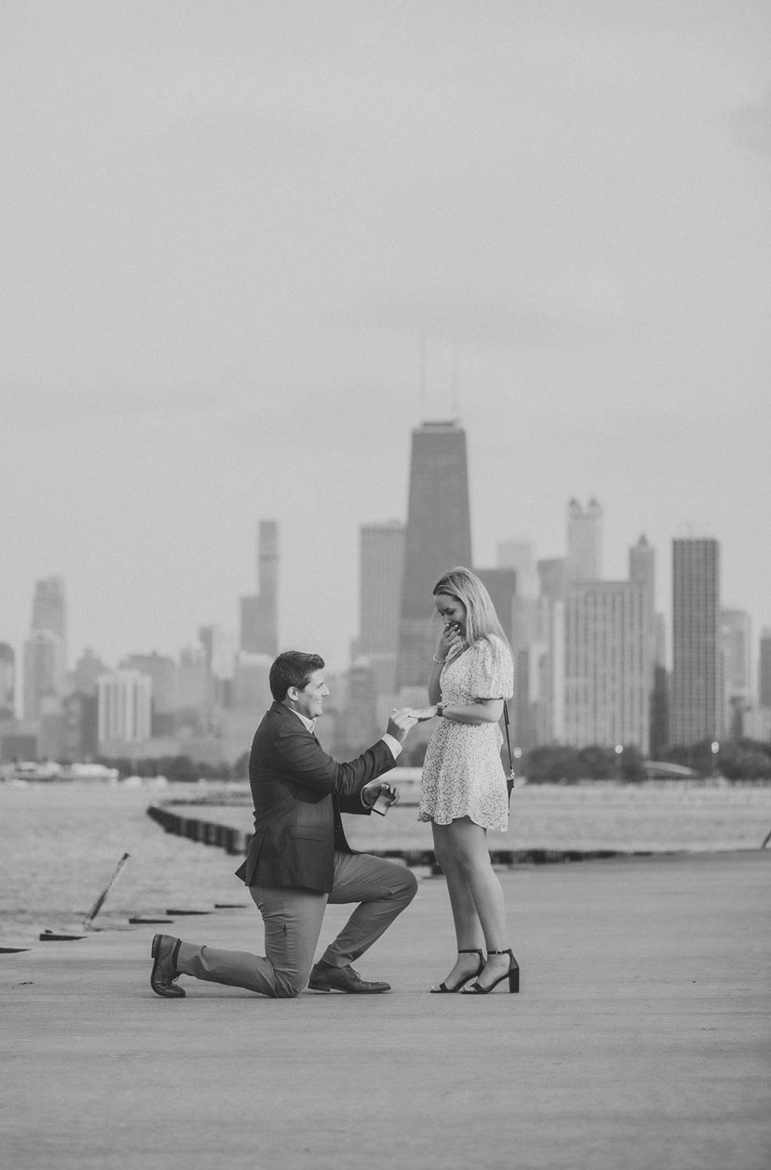 A man proposing to a woman by the waterfront with a city skyline in the background, in black and white.