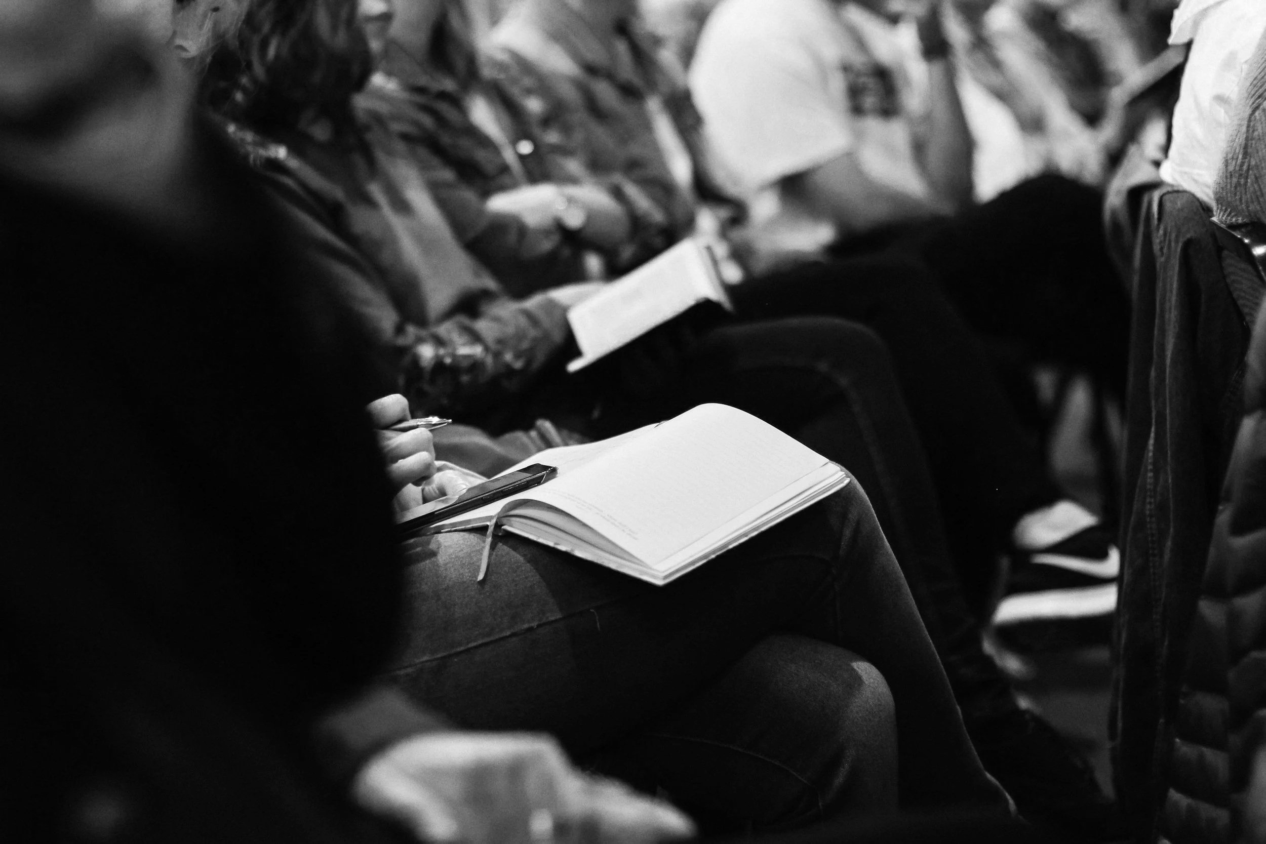 People sitting in an audience, taking notes and reading notebooks during a meeting or lecture.