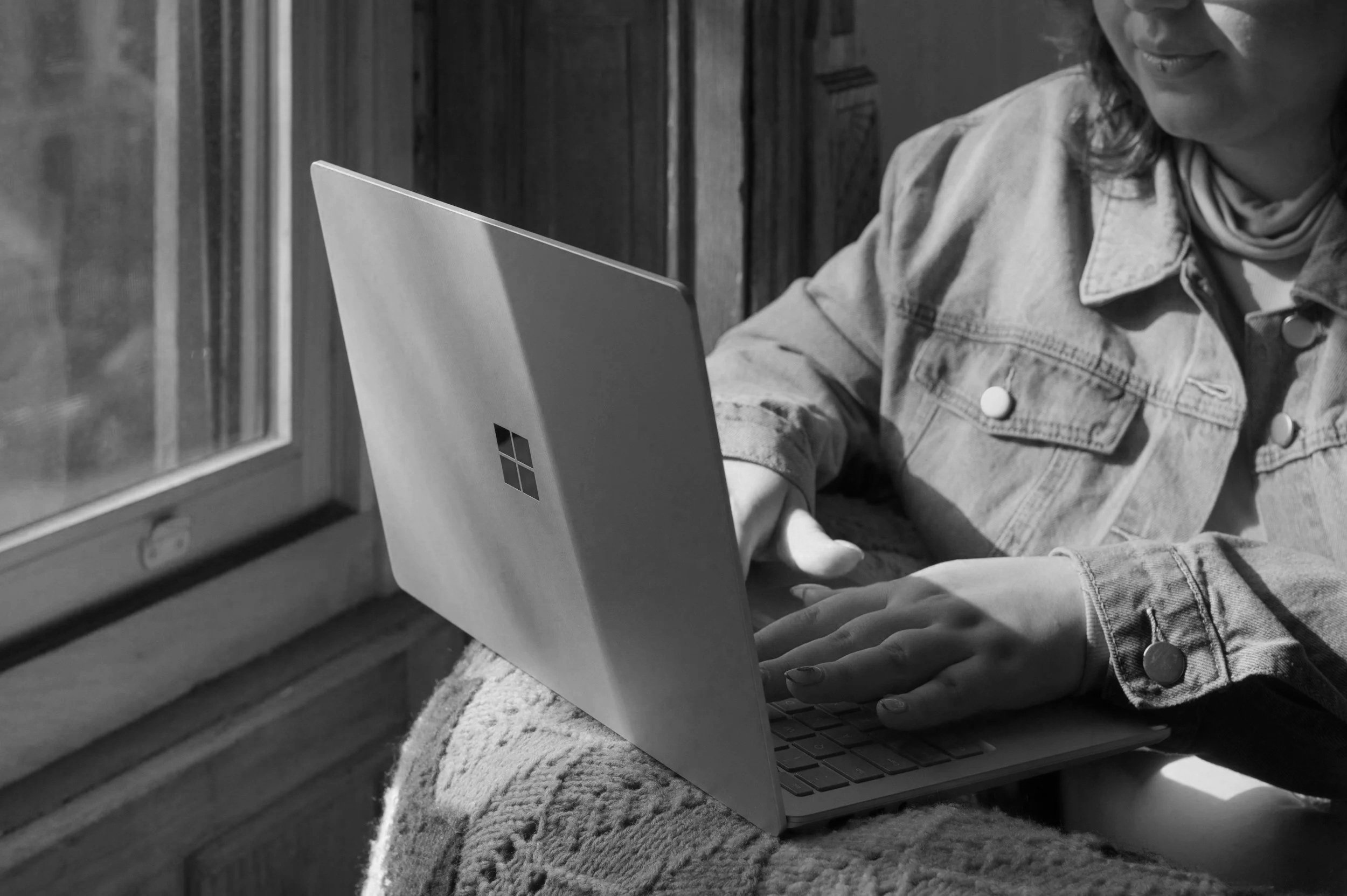A person wearing a denim jacket using a Microsoft Surface laptop near a window.