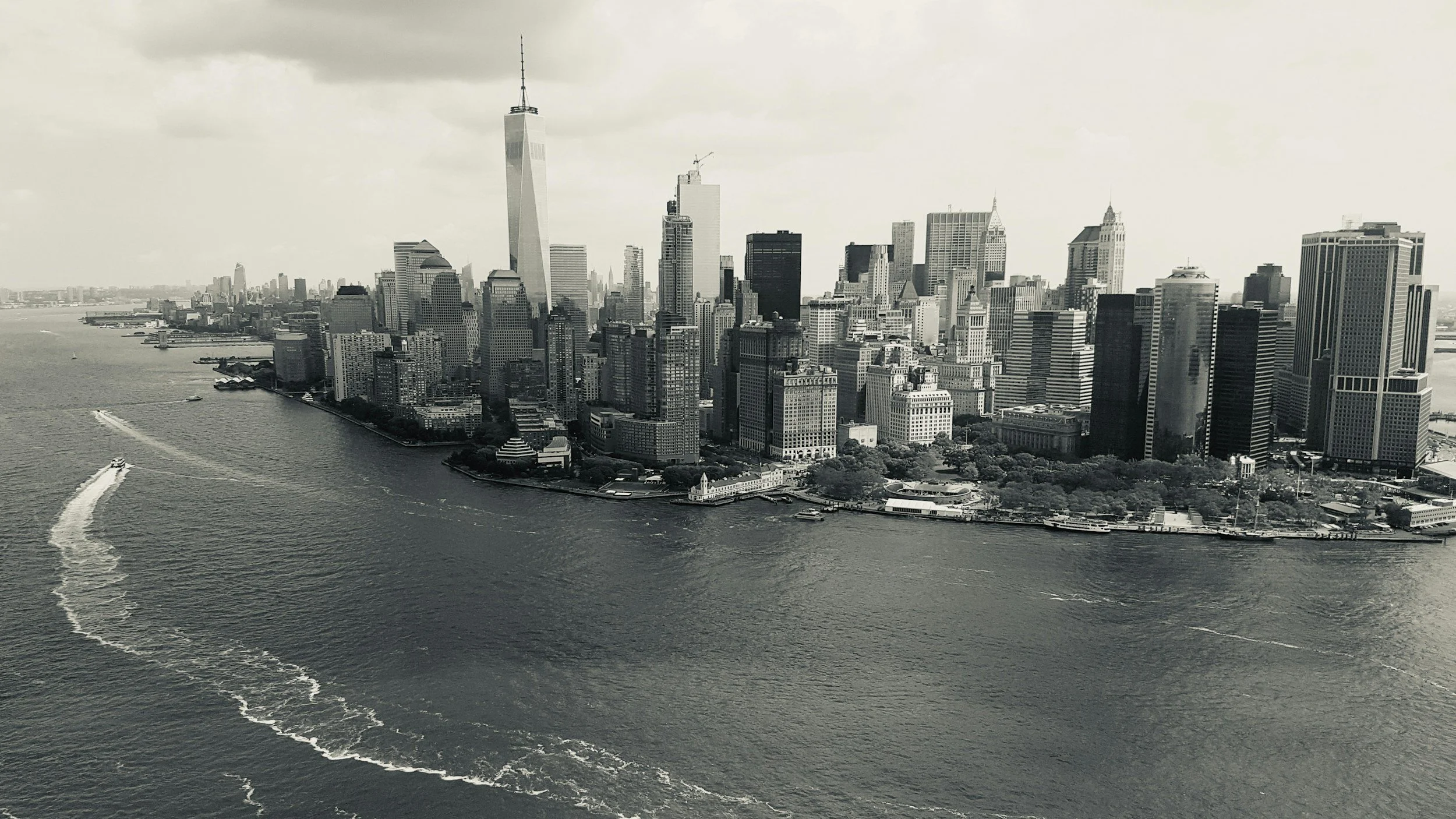 Black and white aerial view of Manhattan skyline with tall skyscrapers along the waterfront, boats on the river, and cloudy sky.