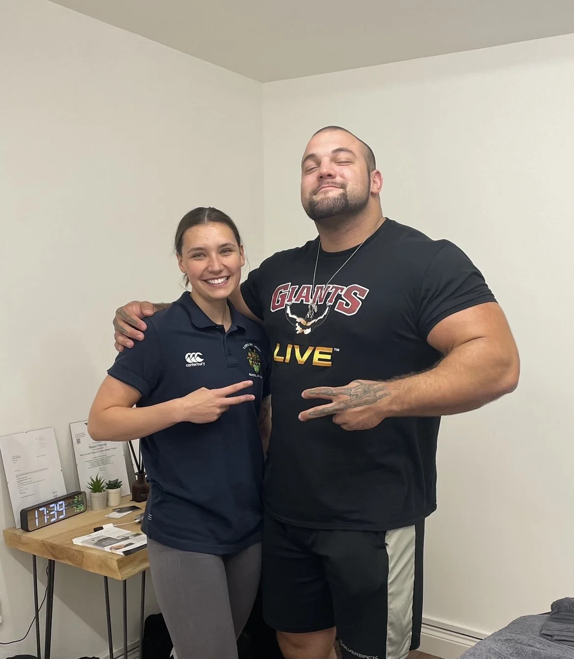 A woman and a muscular man posing in a room, both making peace signs. The woman is smiling and wearing a navy polo shirt, while the man is wearing a black "Giants Live" t-shirt.