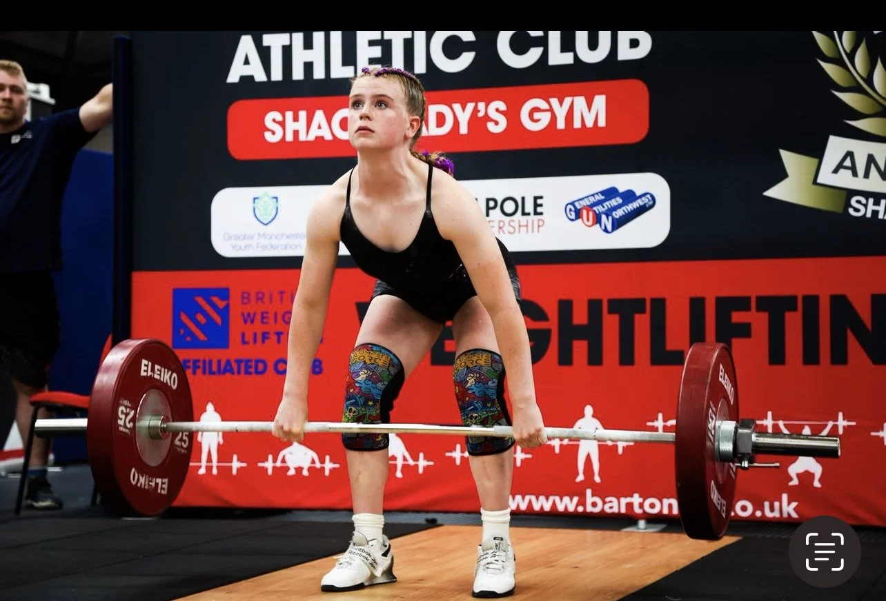 A young woman lifting a barbell during a weightlifting competition at an athletic club, with event banners in the background.