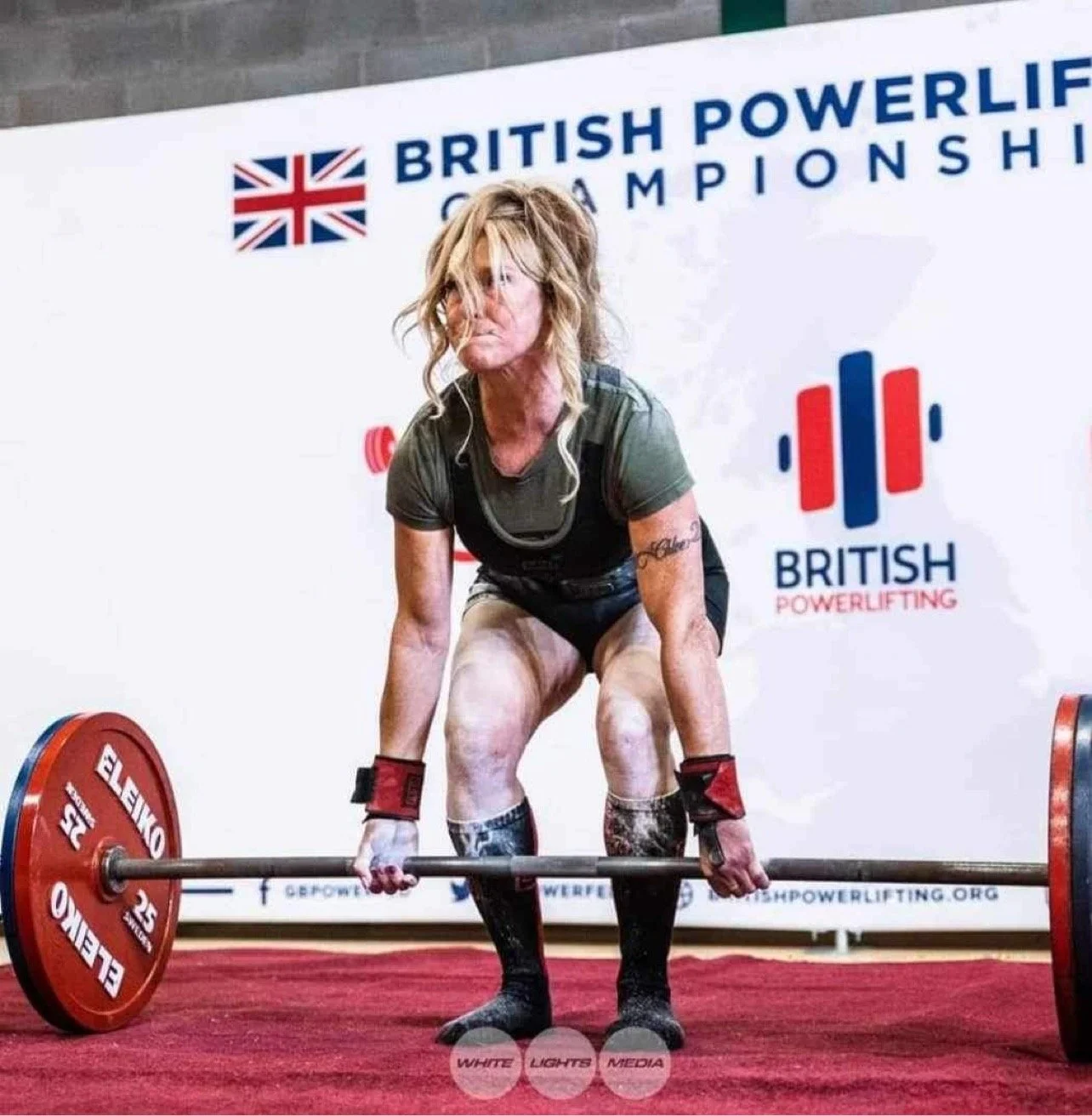 A woman with blonde hair lifting a barbell at a British Powerlifting Championship event.