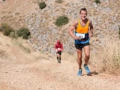 A male runner in a yellow tank top and blue shorts running on a dirt trail in a hilly, desert landscape, with a woman in a red jacket and black shorts running behind him.