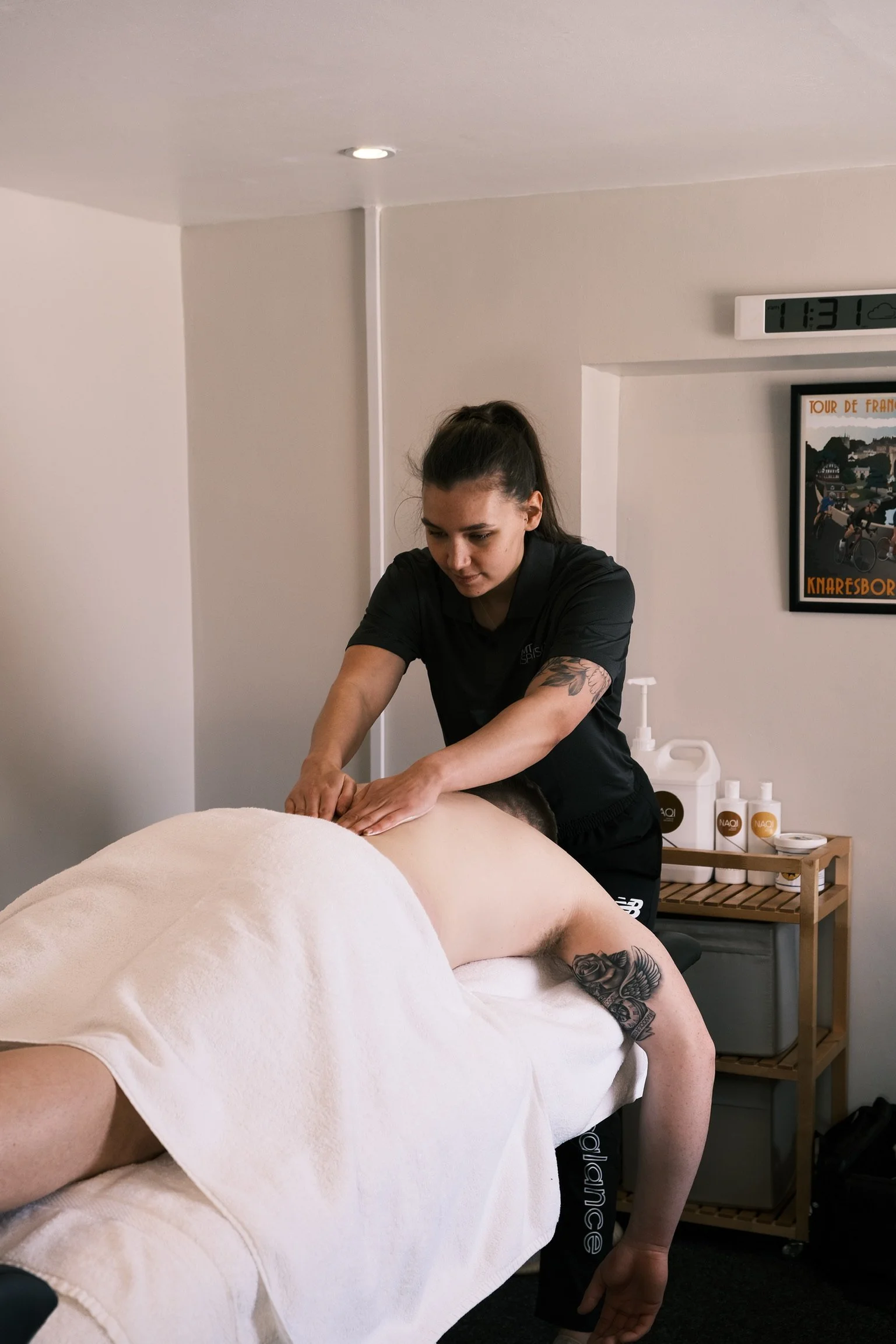 Our practitioner, Megan, giving a sports massage to a person lying face down on a massage table, in a clinic room with a clock and framed picture of the Tour Du Yorkshire on the wall.