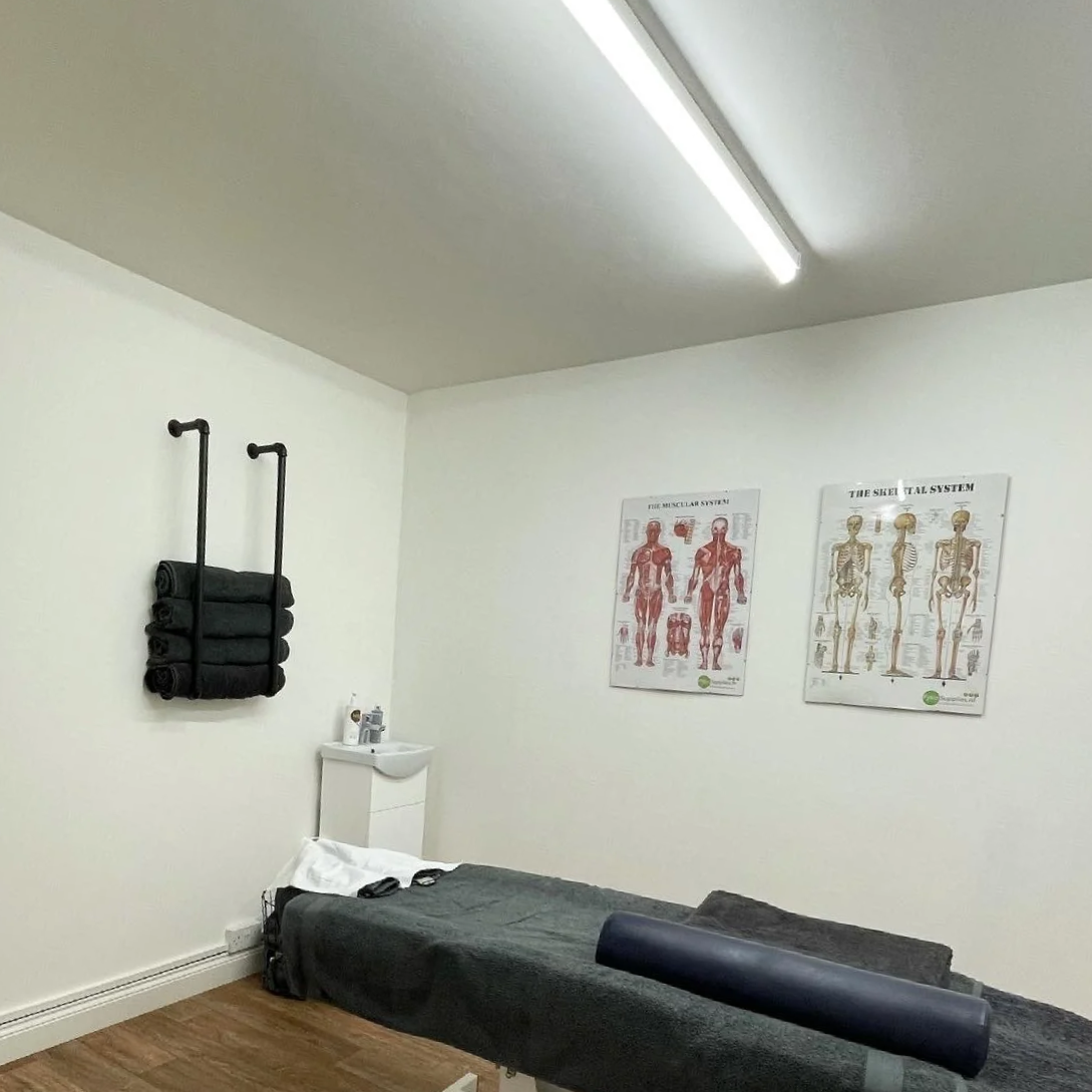 Massage or therapy room with a massage table covered in dark towels, two chiropractic or therapy posters on the wall, black rolled towels hanging on a rack, a small white cabinet with bottles and supplies, and a ceiling light fixture.