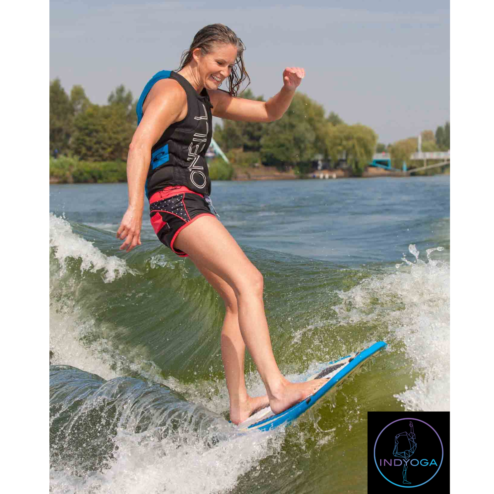 A woman with wet hair wearing a black and blue life vest and red shorts surfing on a wave in a river or lake, smiling and balancing on a blue surfboard with trees and a bridge in the background.