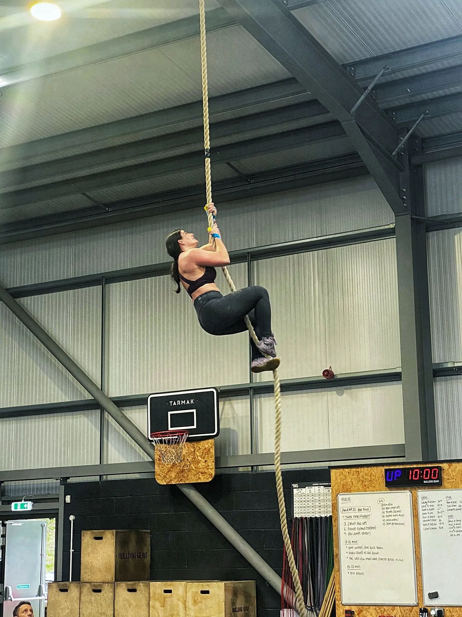 A woman climbing a thick rope inside a gymnasium, with a basketball hoop and scoreboard visible in the background.