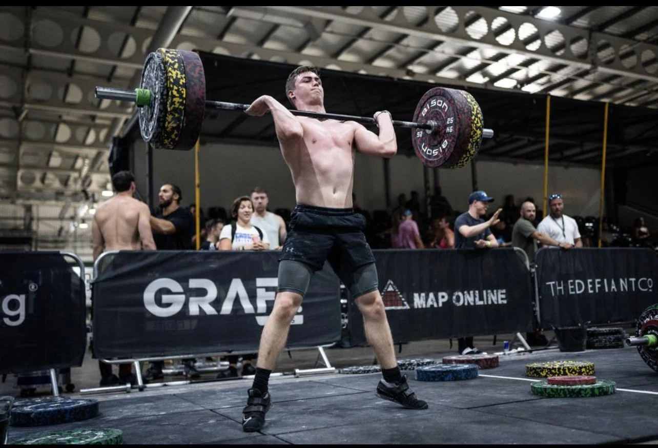 A male weightlifter performing a clean and jerk at a CrossFit competition, lifting a loaded barbell overhead.