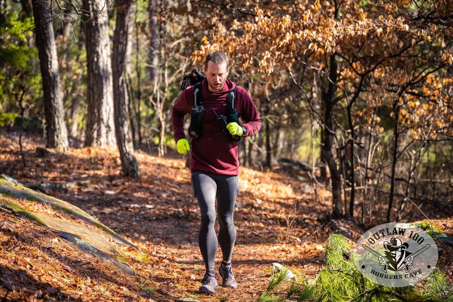 A man running through a wooded trail with autumn leaves on the ground, wearing a maroon sweatshirt, black running tights, and bright yellow gloves, carrying a backpack.