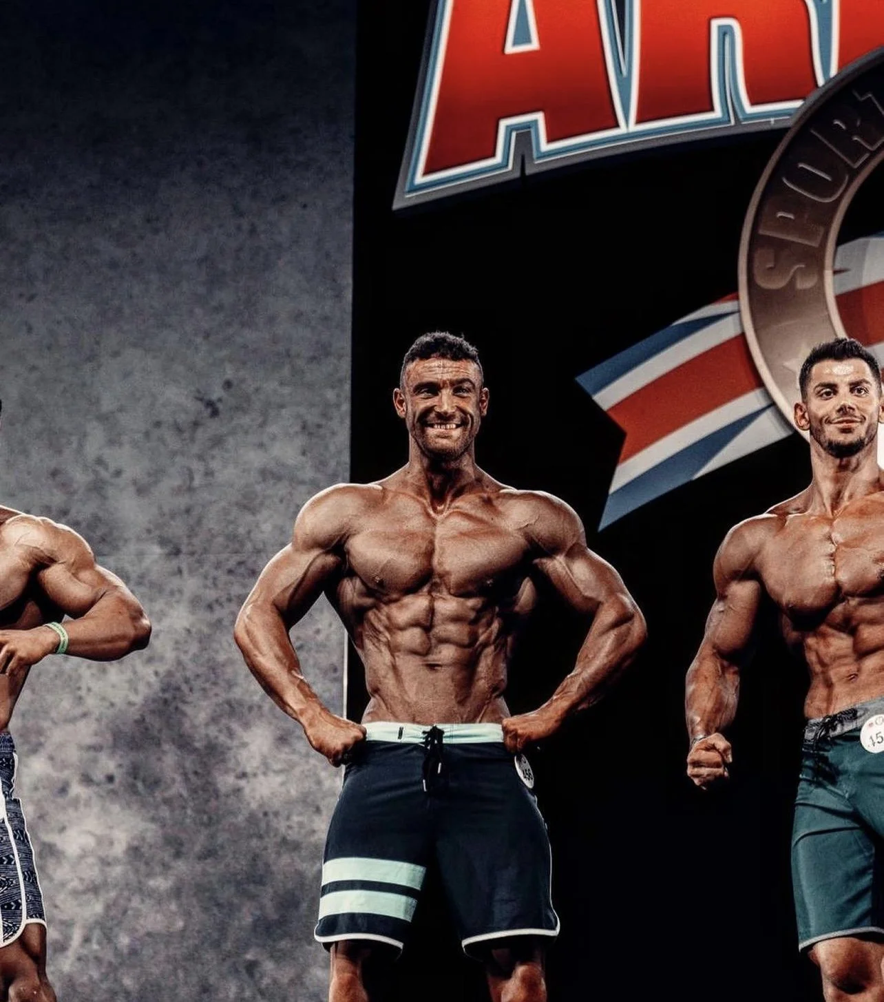 Three male bodybuilders on stage at a competition, flexing and posing shirtless with visible muscles, with a logo or banner in the background.