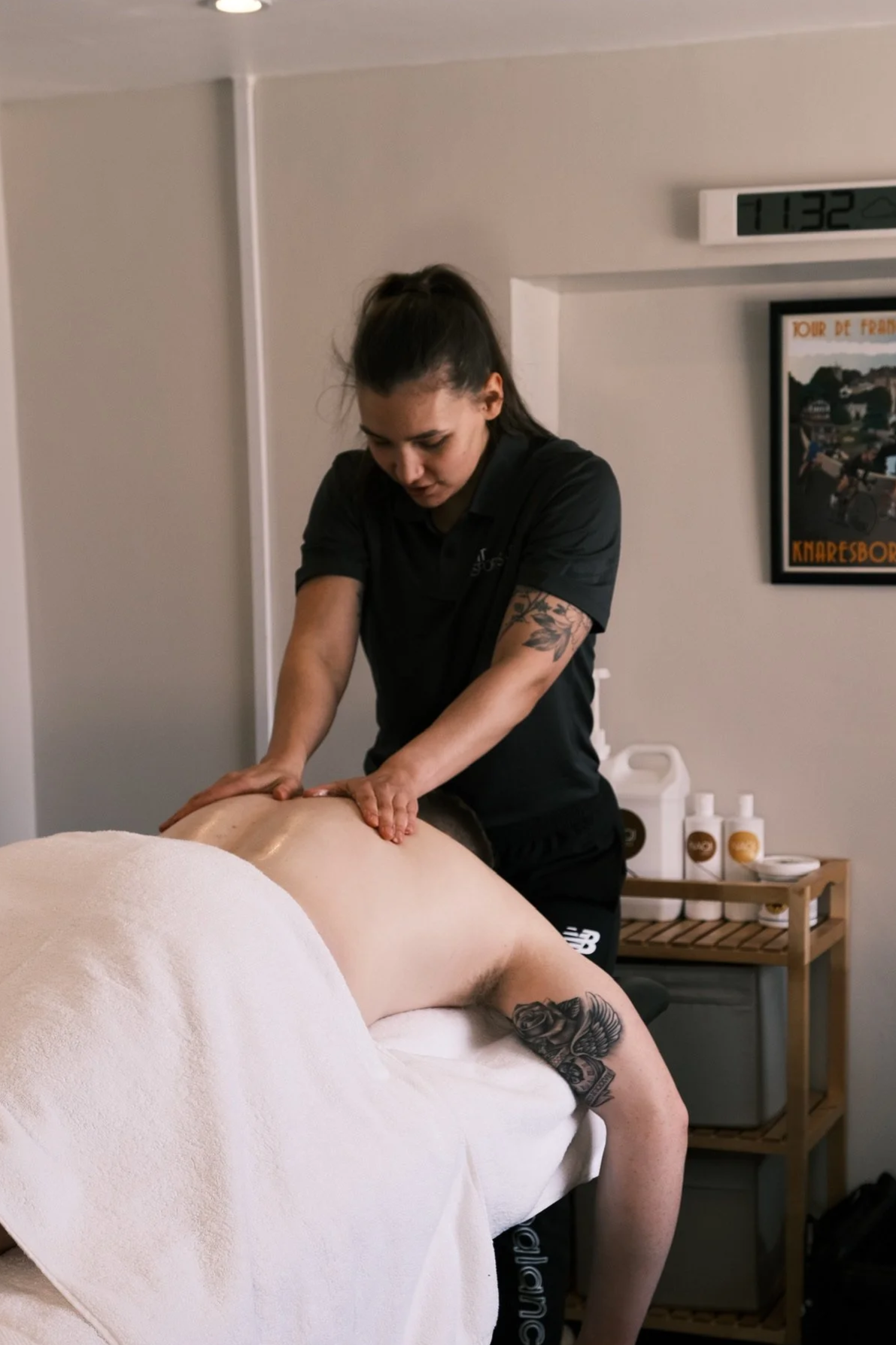 A massage therapist performing a massage on a person lying face down on a massage table, in a therapy room with a clock and framed map on the wall.