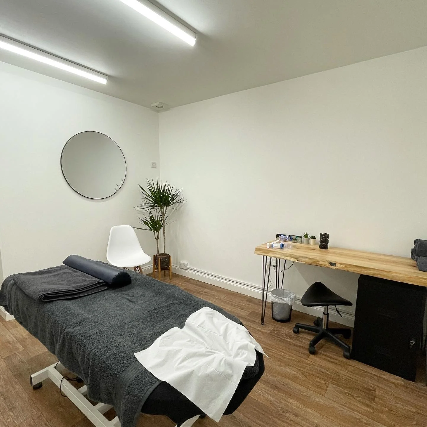A massage therapy room with a massage table covered in dark gray towels, a rolled towel or bolster, and a white cloth at the foot of the table. There's a white chair near a tall potted plant in the corner, a round mirror on the wall, and a wooden desk with massage oils, lotions, and small plants. A black rolling stool and a small black cabinet are under the desk, with a trash bin nearby. The room has hardwood floors and fluorescent ceiling lights.