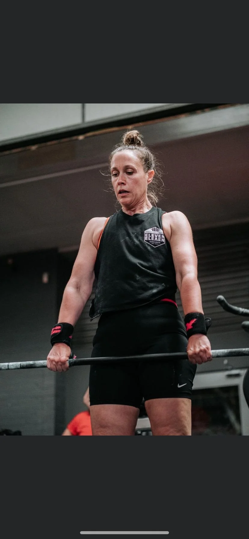 A woman lifting a barbell in a gym, wearing black workout clothes and wristbands.