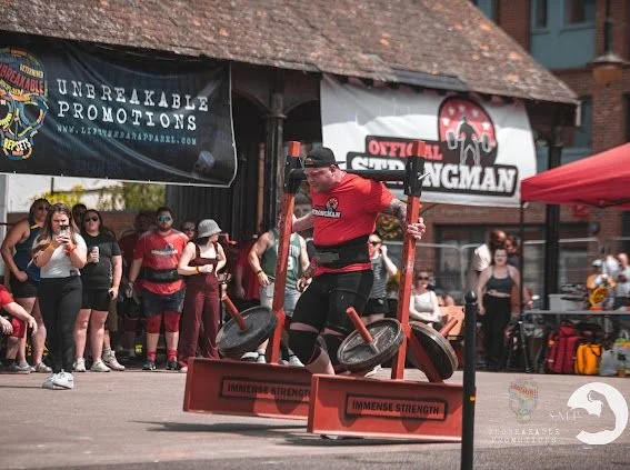 A strong man participating in a tire-flipping competition at an outdoor event, surrounded by spectators and event banners.