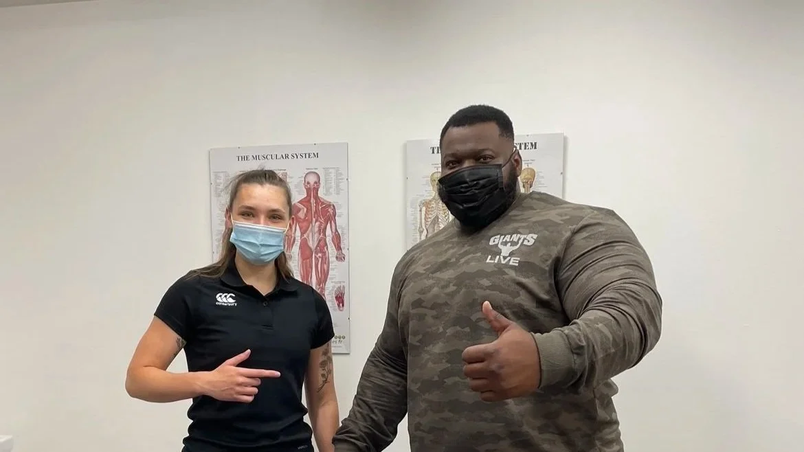 A woman and a man wearing face masks, standing in front of anatomical posters, giving thumbs up and pointing at each other.