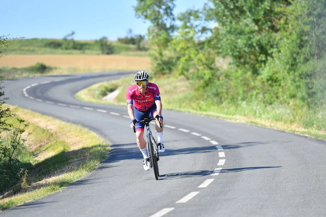 A cyclist riding a road bike on a winding country road surrounded by green trees and grass.