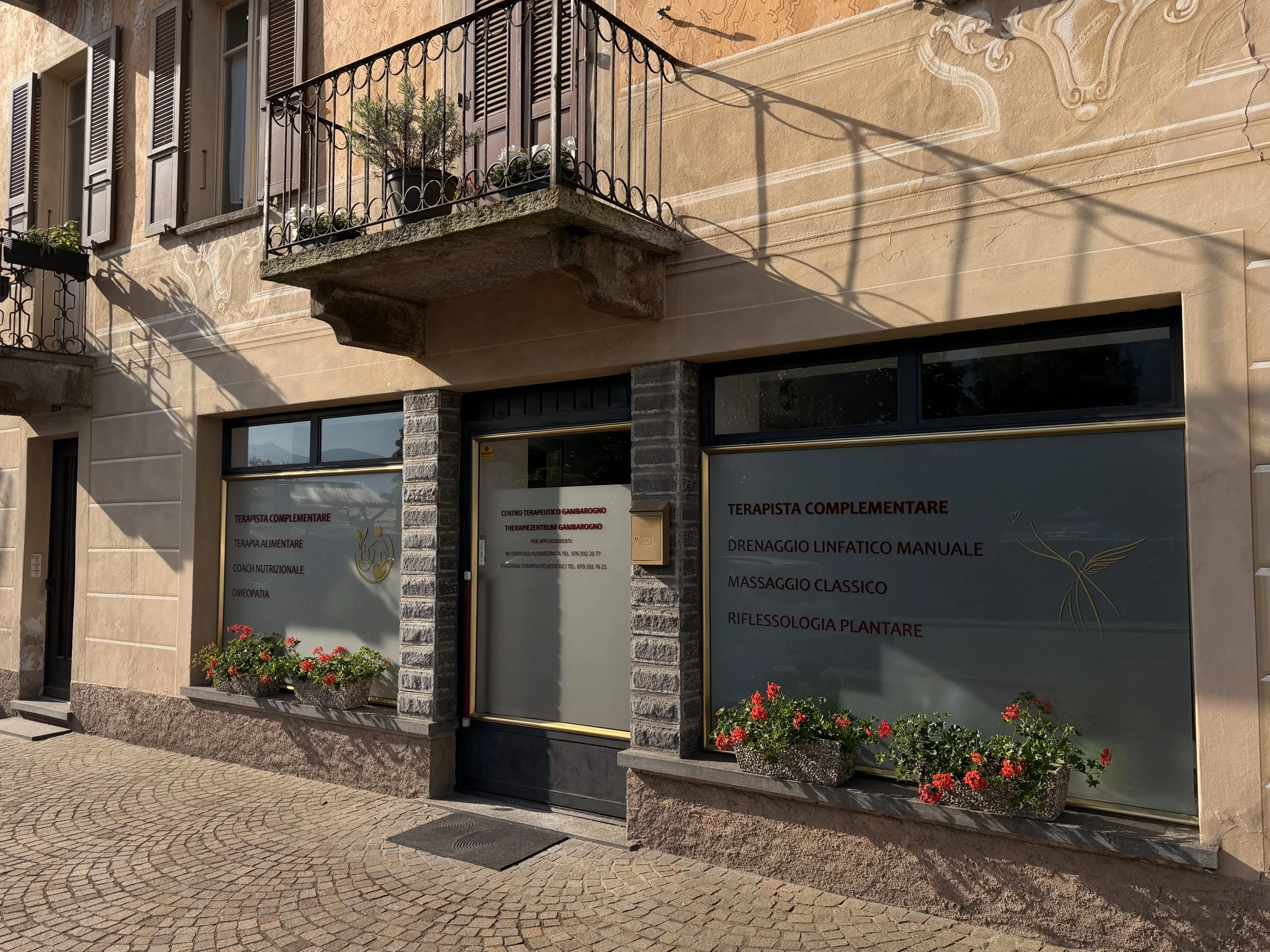 Vetrina di un centro terapeutico con insegne che indicano servizi come terapia complementare, terapia alimentare, coaching nutrizionale, omeopatia, drenaggio linfatico manuale, massaggio classico, riflessologia plantare. La vetrina è decorata con fioriere piene di gerani rossi e un balcone con piante. L'edificio ha pareti color ocra e dettagli in pietra e metallo.