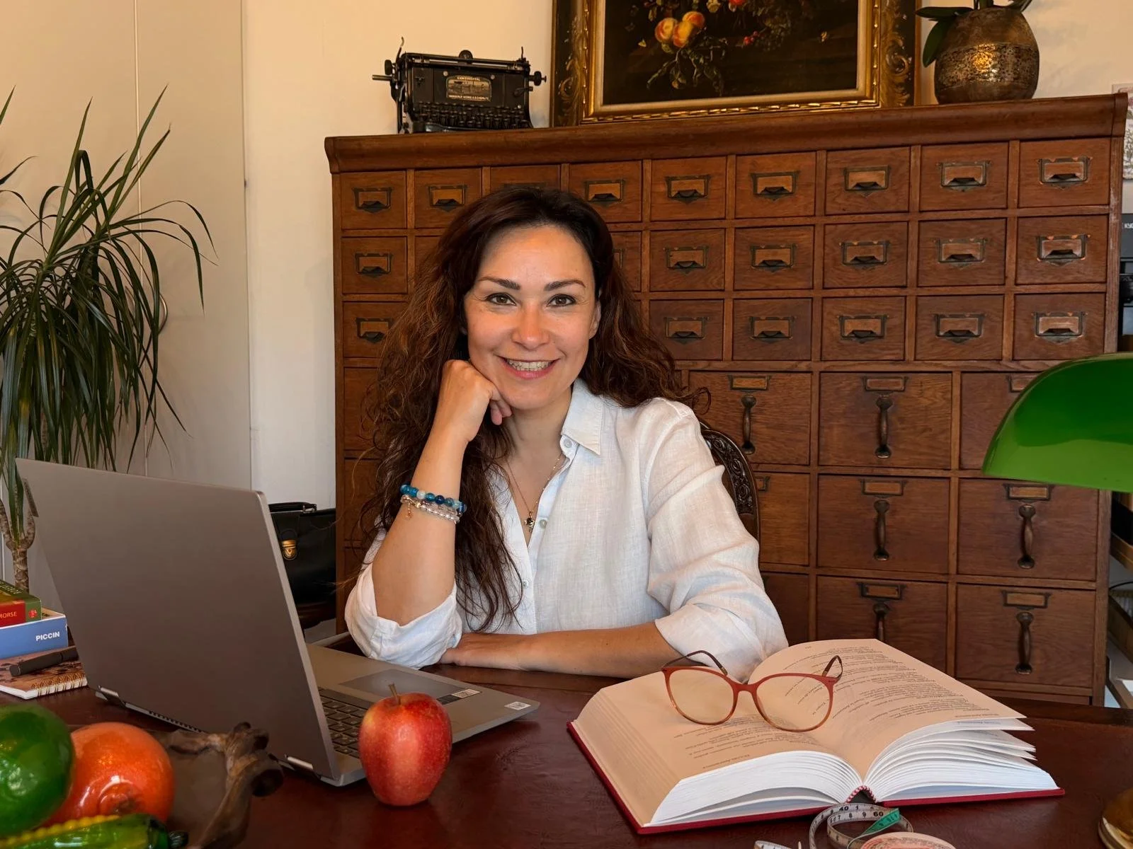 Donna sorridente seduta a una scrivania con libro aperto, taccuino, occhiali rossi e laptop, in uno studio arredato con mobili in legno e piante verdi.