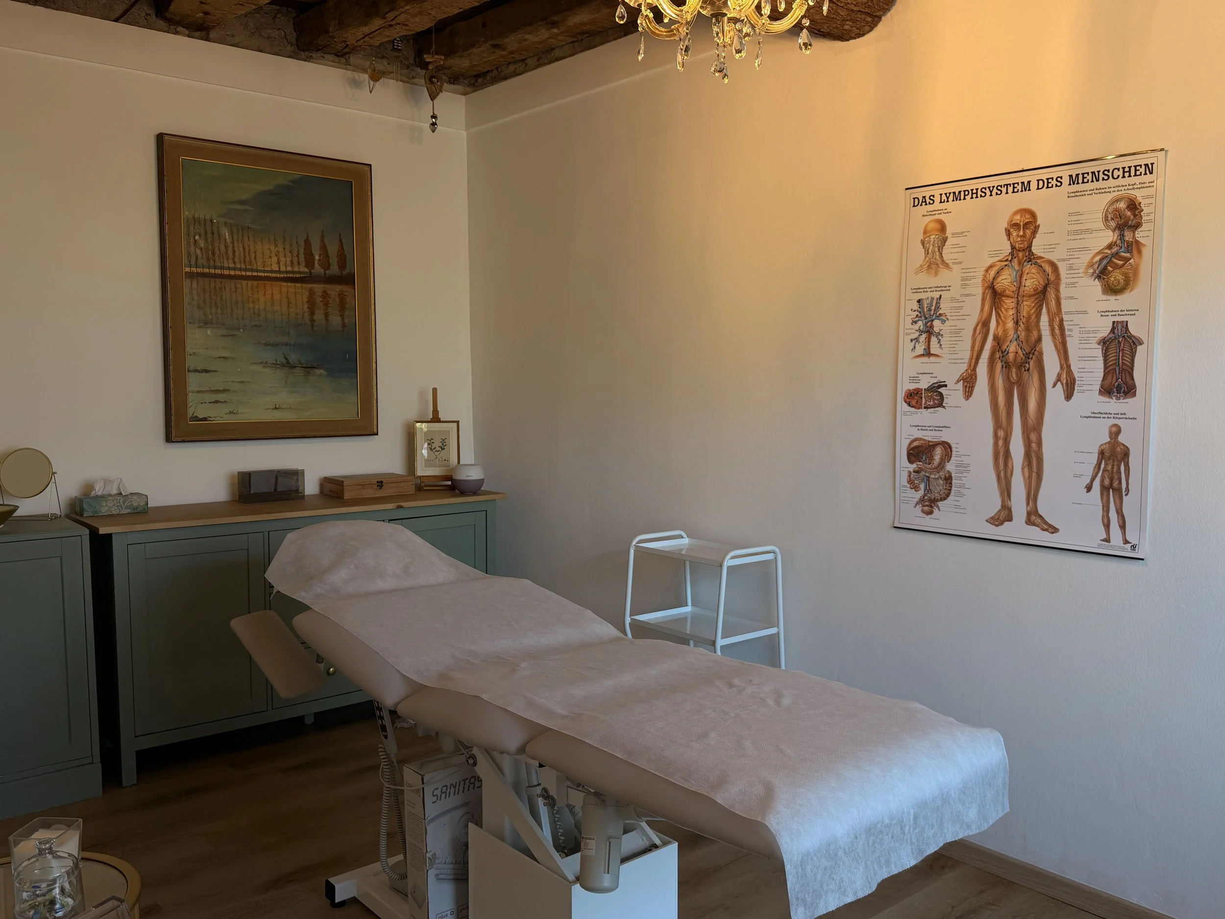 Una stanza di uno studio medico con un letto per trattamenti, un armadietto e un quadro sull'istitologia del sistema linfatico nel muro.