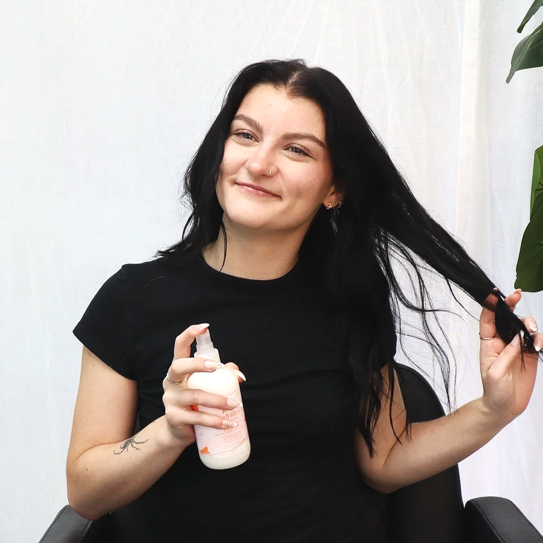 A woman with black hair and a nose ring smiling while holding a bottle of hair product in one hand and brushing her hair with the other.