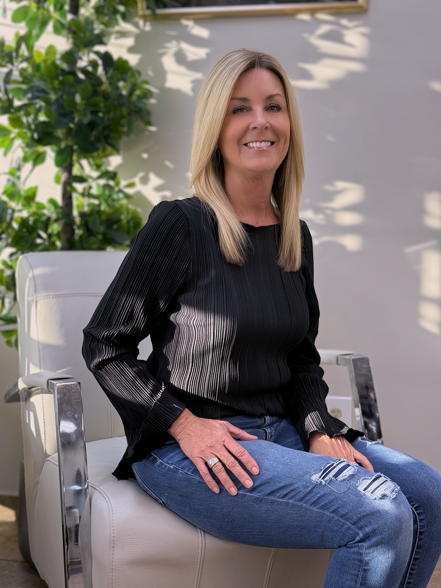 A smiling blonde woman sitting on a white chair outdoors, wearing a black pleated blouse and ripped jeans, with green plants behind her.