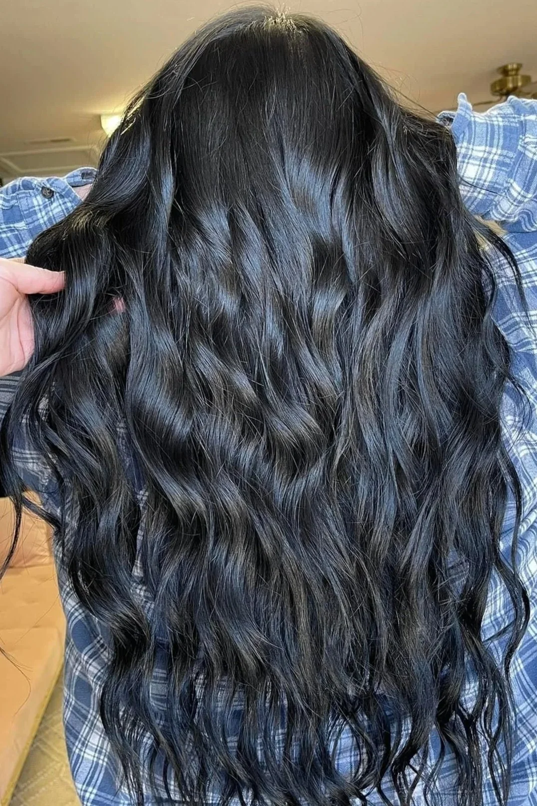 Long, dark, wavy hair on a person wearing a blue plaid shirt, with a hand holding a section of the hair.