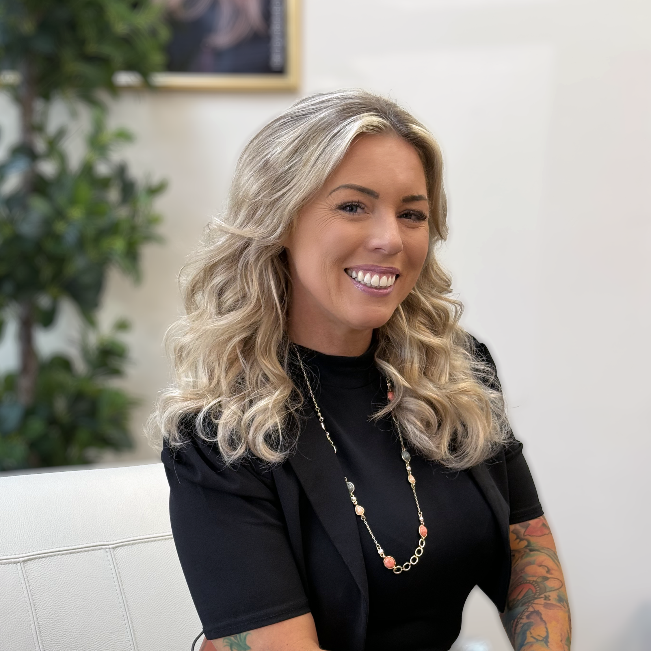 A smiling woman with wavy blonde hair, wearing a black top and a long necklace, sitting indoors in front of a white wall with a framed picture and a potted plant in the background.