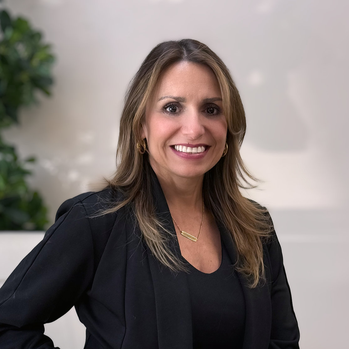 A smiling woman with brown hair and gold jewelry, wearing a black blazer, standing indoors with a blurry plant in the background.