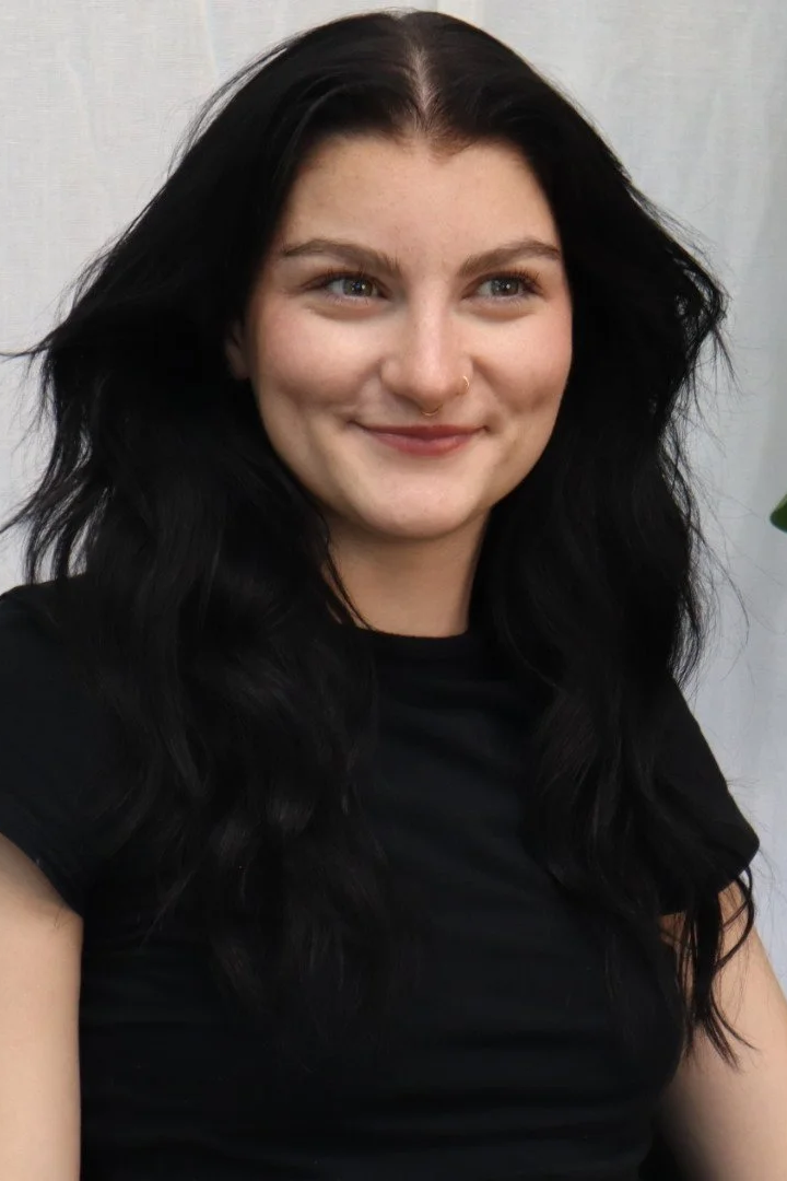 A young woman with long black wavy hair, light skin, and a septum piercing, smiling and looking to the side, wearing a black shirt, in an indoor setting.