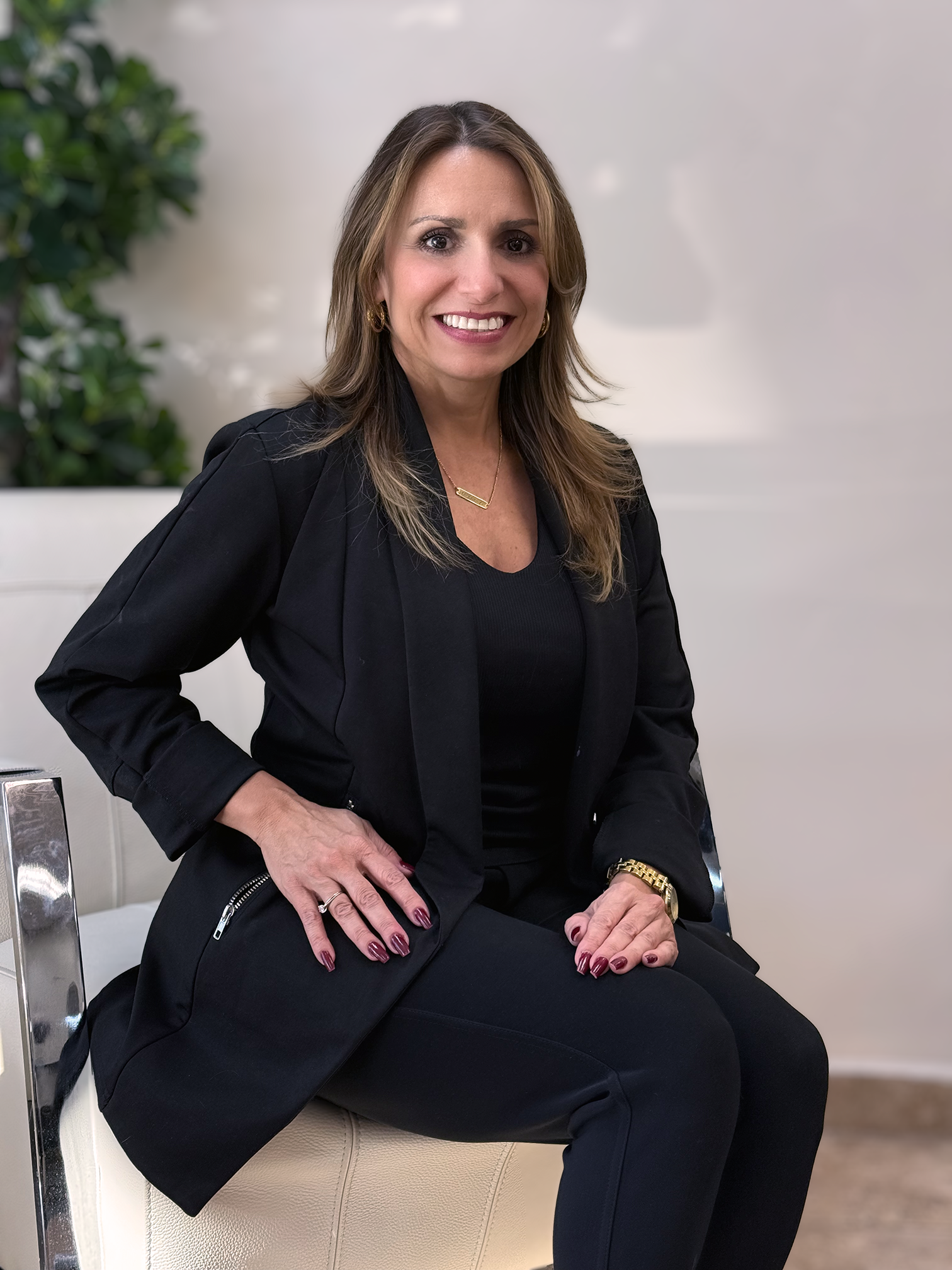 A woman sitting on a beige chair, wearing a black blazer and pants, with long light brown hair, smiling, with plants and a white wall in the background.