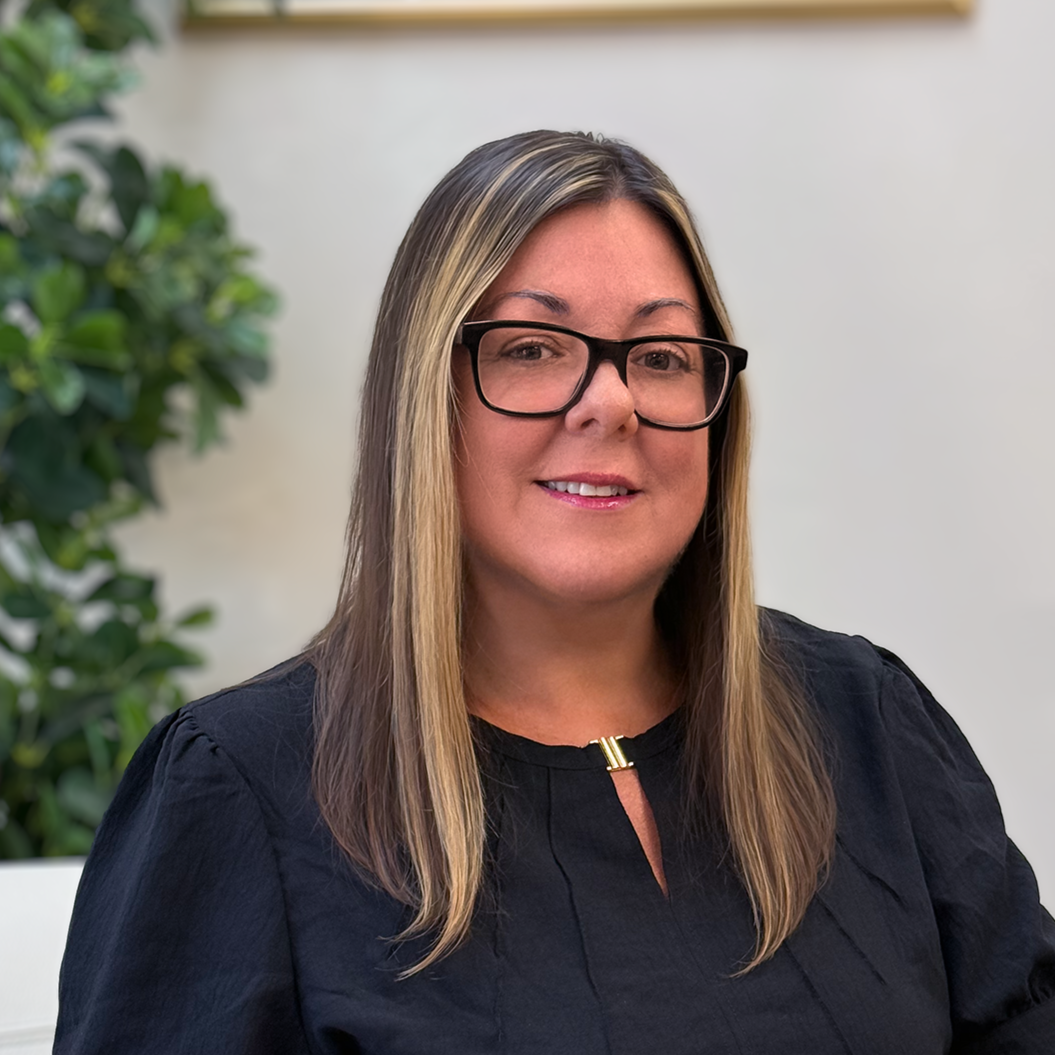 A woman with shoulder-length blonde hair, wearing black glasses and a black top, smiling indoors with a plant in the background.