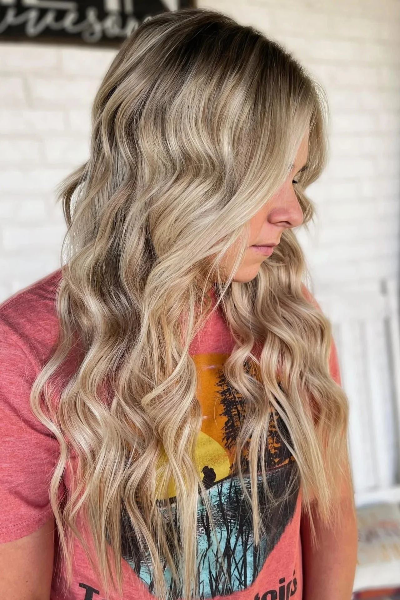 Side profile of a woman with long, wavy blonde hair, wearing a pink T-shirt with a colorful sunset landscape graphic.