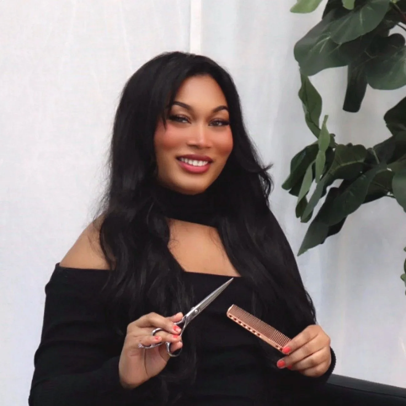 A woman with long black hair, wearing a black top, holding hair cutting scissors and a hair comb, smiling at the camera, standing beside a green plant against a white wall.