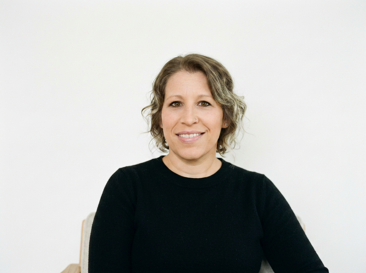 A woman with shoulder-length curly hair wearing a black shirt, smiling, sitting against a plain white background.