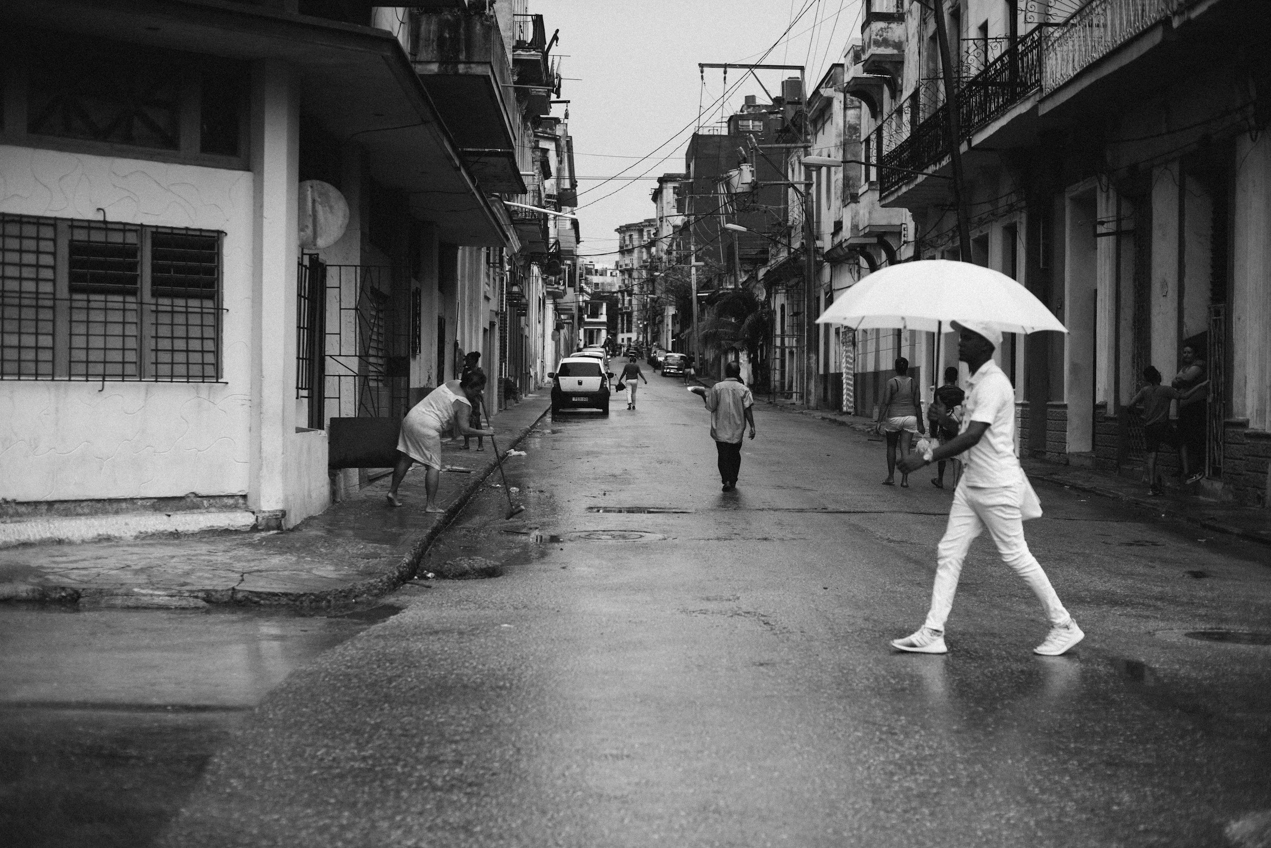 Una calle urbana en un día lluvioso con varias personas caminando, algunos con paraguas y otros encharcando, en un entorno de edificios residenciales.