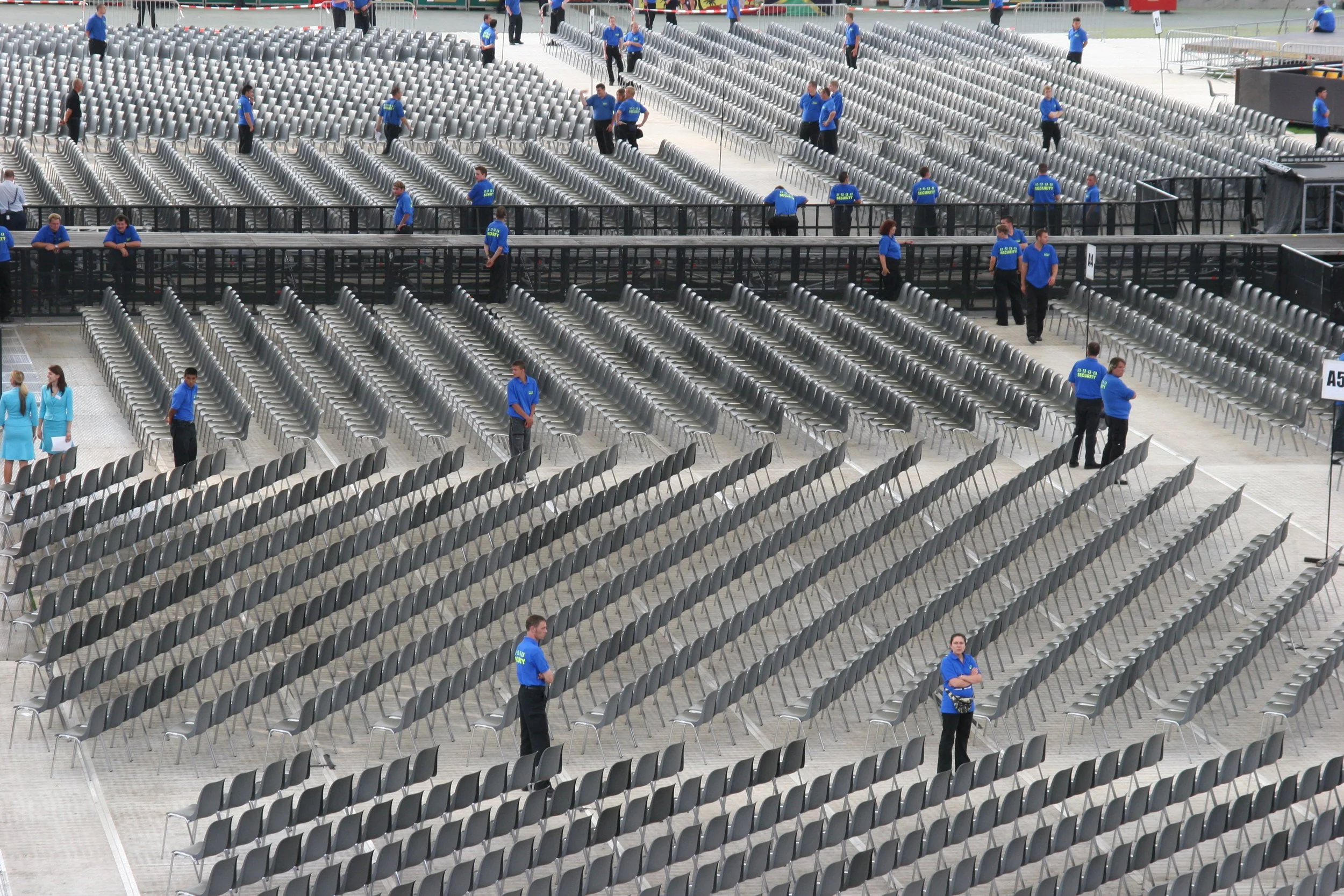 Leere Sitzreihen bei einer Veranstaltung mit mehreren Personen, die Sicherheitsdienste sind. Tische und Stühle sind aufgestellt, das Gebäude ist modern gestaltet.